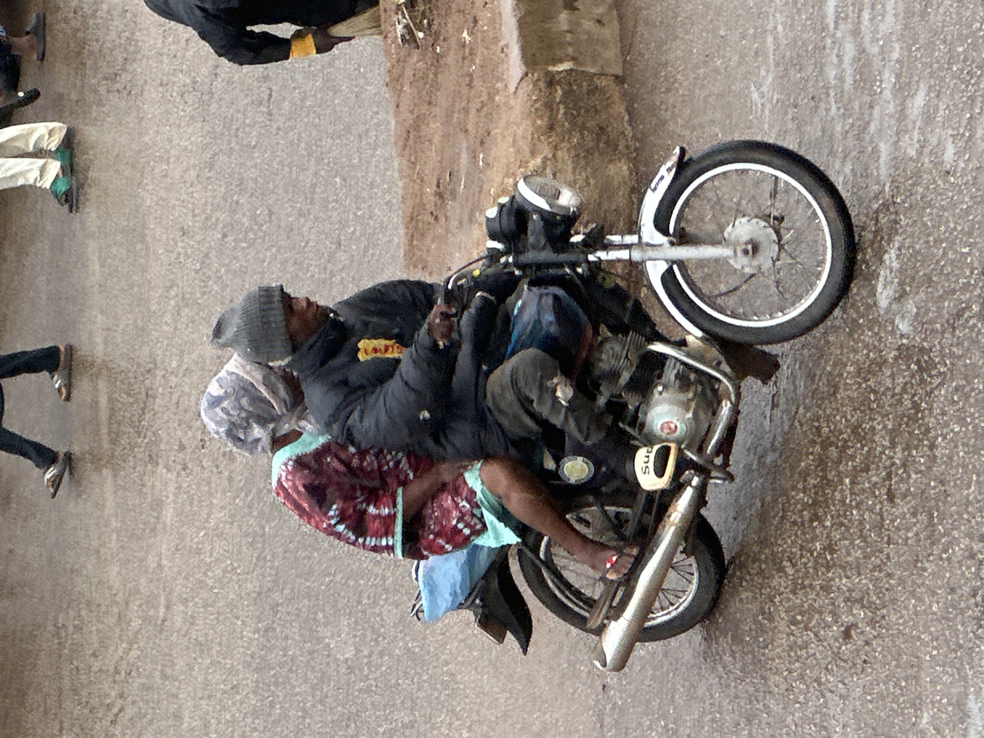 Two people are riding on a motorcycle on a paved surface, likely a road or street. The person in front is wearing a dark jacket and a grey beanie, and appears to be driving. The person behind is wearing a patterned garment, possibly a dress or wrap, and a headscarf. Their legs are visible, and they are wearing sandals. The motorcycle is a mid-sized model with a silver exhaust pipe. The scene is outdoors, and the lighting suggests it could be daytime, possibly overcast or late afternoon. The ground surface appears to be rough asphalt or concrete, with some scattered debris. The overall impression is one of everyday life in a possibly busy urban or semi-urban environment. There is some text visible on the motorcycle, which appears to be a logo or brand name, possibly starting with "Sup". The people's postures and expressions are not clearly discernible due to the angle and resolution of the image, but the activity of riding a motorcycle together is evident.