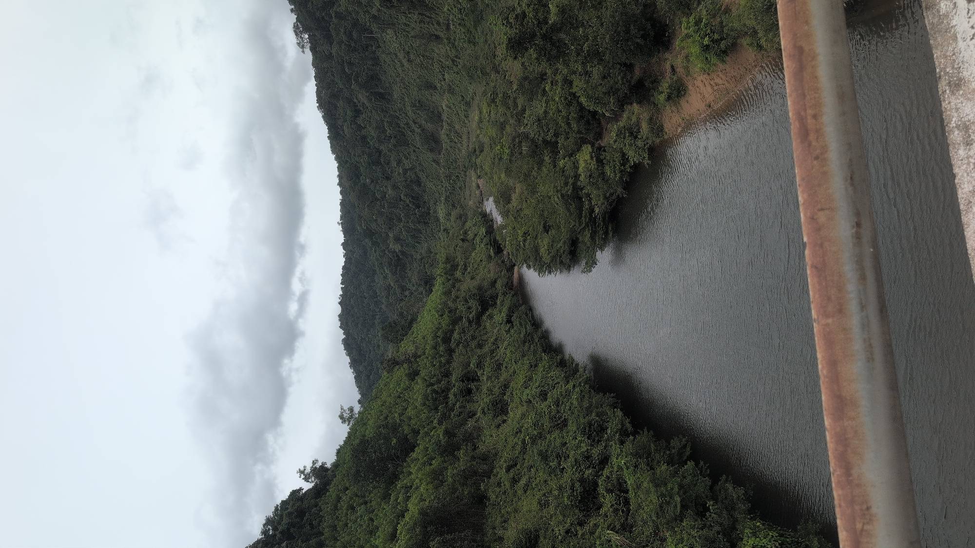 The image shows a serene river flowing through a lush green valley. Dense foliage covers the rolling hills on either side, with a cloudy sky overhead. In the foreground, a rusty metal railing runs horizontally across the frame, suggesting the viewpoint is from a bridge. The river, with its dark, rippling water, winds its way into the distance, disappearing among the trees. The scene evokes a sense of natural tranquility, likely captured in a rural or mountainous area. The location is identified as Trường Sơn Commune, Vietnam.