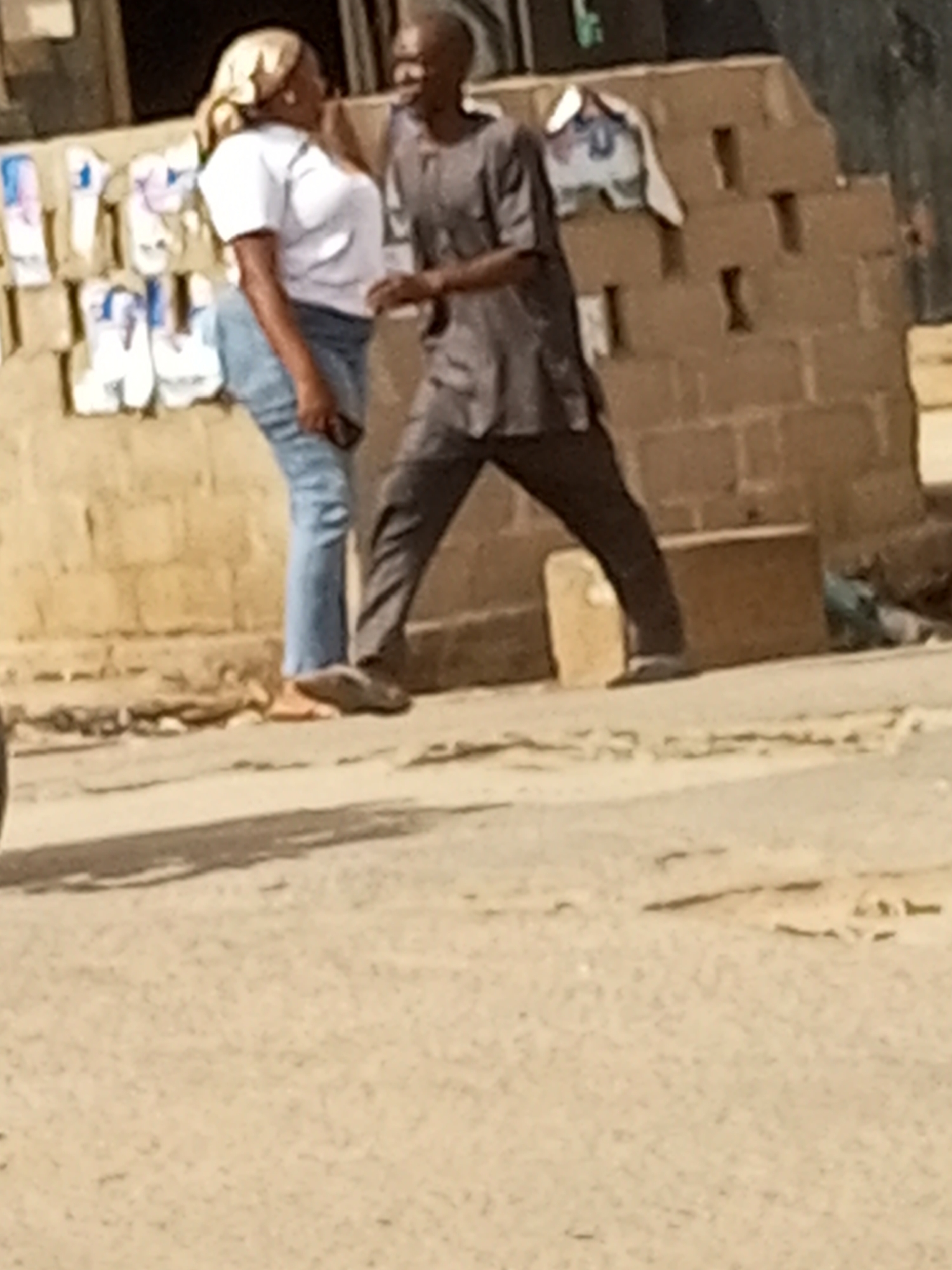 Two people, a woman and a man, are standing on a dusty street in Maiburiji, Nigeria. The woman is wearing a white t-shirt, blue jeans, and a headscarf. The man is wearing a grey dashiki and dark pants. They appear to be talking to each other. In the background, there is a low brick wall, on which hang items that appear to be for sale. Further back, there is a building, constructed from dark, roughly hewn bricks. The foreground consists of a dusty road.