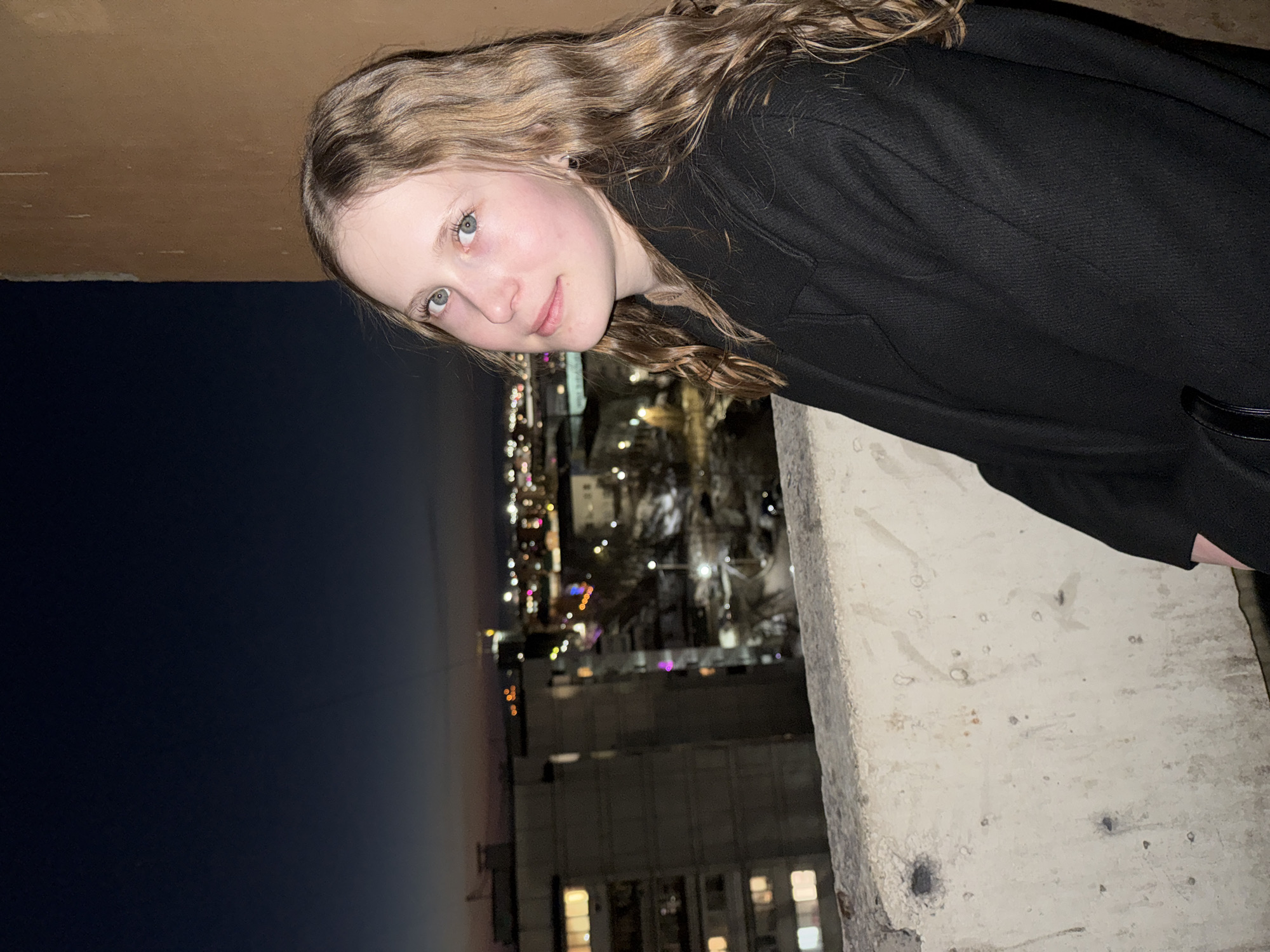 A young woman with long, wavy blonde hair and light eyes, wearing a black coat, leans out from a concrete structure overlooking a city at night. The city lights are visible in the background, suggesting a cityscape. The location is likely Chaikovsky, Russia. The image is taken from a low angle, emphasizing the height from which the woman is leaning. The scene depicts a person interacting with an urban environment at night.