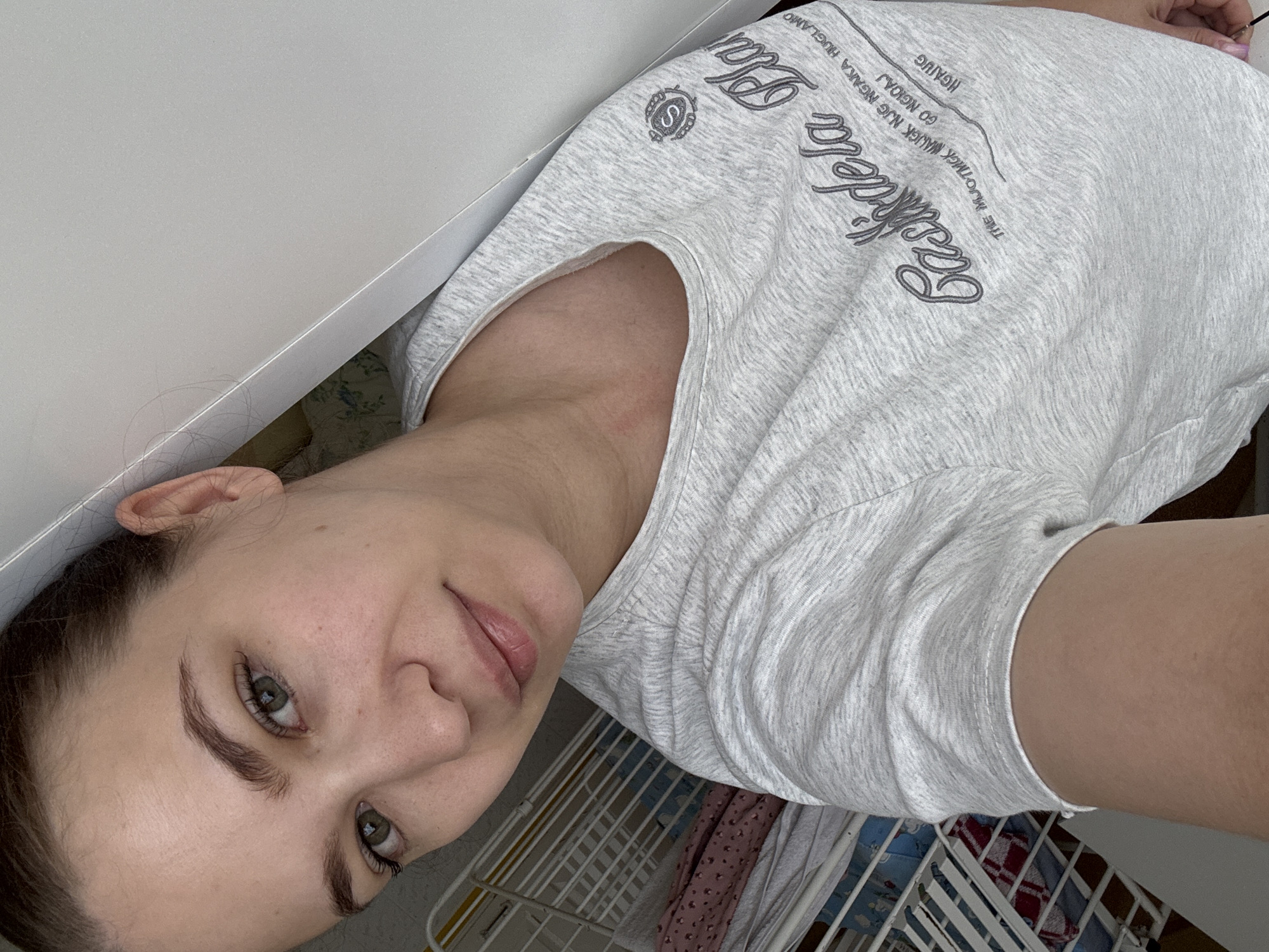 A young adult female is depicted in a close-up, selfie-style photograph, gazing directly into the camera with a neutral to slight smile. Her hair is pulled back, revealing her ears and framing her face. She has fair skin, pronounced dark eyebrows, and green eyes with visible eyelashes. Minor blemishes or freckles are present on her cheeks and under her eyes. Her lips appear natural, with a light pink hue. She is wearing a light gray heathered short-sleeved t-shirt with a rounded neckline. The t-shirt features dark gray text on the front, including "Cashidela Pla" in a script font, and smaller capitalized text below: "THE MUOTIMGK MNUEK NJG NGAKA HUGLAMO GO NGIOAJ" followed by "NGAUG". A circular logo containing an "S" within a crest or shield design is located on the upper right chest of the shirt.
The background to the viewer's left includes a white wire shelving unit, behind which various folded textiles are visible, including a pink patterned fabric and white material. To the viewer's right and in the upper portion of the image, a plain white, matte surface, likely a wall or furniture panel (such as a wardrobe door), dominates the frame. A thin, light-colored edge or seam runs diagonally across this white surface. Behind a slight gap in the white surface, a glimpse of a light-colored fabric with a floral pattern is visible. The scene is situated indoors, specifically within a residential or private setting located in the city of Chaikovsky, Russia.