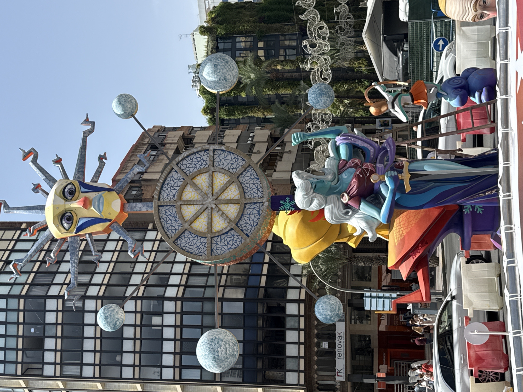 In a city setting, likely a public square or street, a large, elaborate artistic sculpture is the central focus. The sculpture appears to be part of a festival or celebration, possibly related to astronomy or mythology, given the prominent sun-like face at the top and spherical celestial bodies suspended around it.
The main figure in the sculpture is a large, blue-skinned humanoid with white hair, wearing a purple and red garment, and holding a sword. To the right of this figure, a smaller sculpture of a person in orange attire, resembling a cupid or an archer, is perched atop a blue, snail-like creature.
The sculpture is situated in front of modern buildings, one of which has a facade covered in greenery. In the background, there are people walking and cars driving on the street. The atmosphere is bright and sunny, suggesting daytime. The sculpture itself is a multi-tiered construction with vibrant colors and intricate details, including a large, circular, wheel-like element with geometric patterns. Barriers and traffic cones are visible in the foreground, indicating a cordoned-off area for the display.