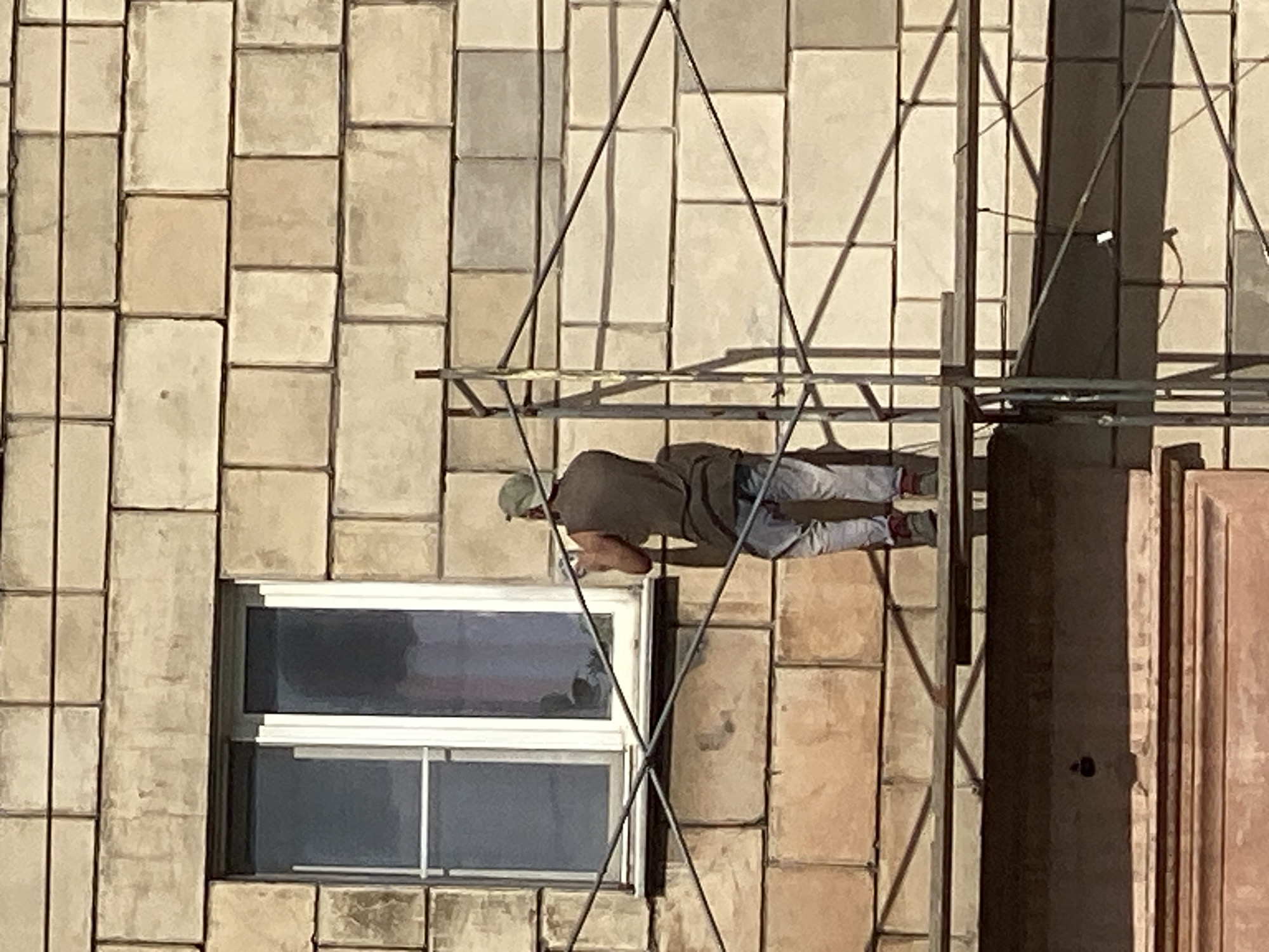 A man in a green cap and a dark sleeveless shirt is standing on scaffolding next to a building in Novokuznetsk, Russia. He is wearing light-colored pants and dark shoes. He is holding a white object in his right hand, possibly a tool or a cleaning item, and appears to be working on the building's facade, which is constructed of large, light-brown stone blocks. A window with white trim is visible to his left. The scaffolding consists of metal poles and horizontal planks. The image is taken from a low angle, looking up at the man and the building.