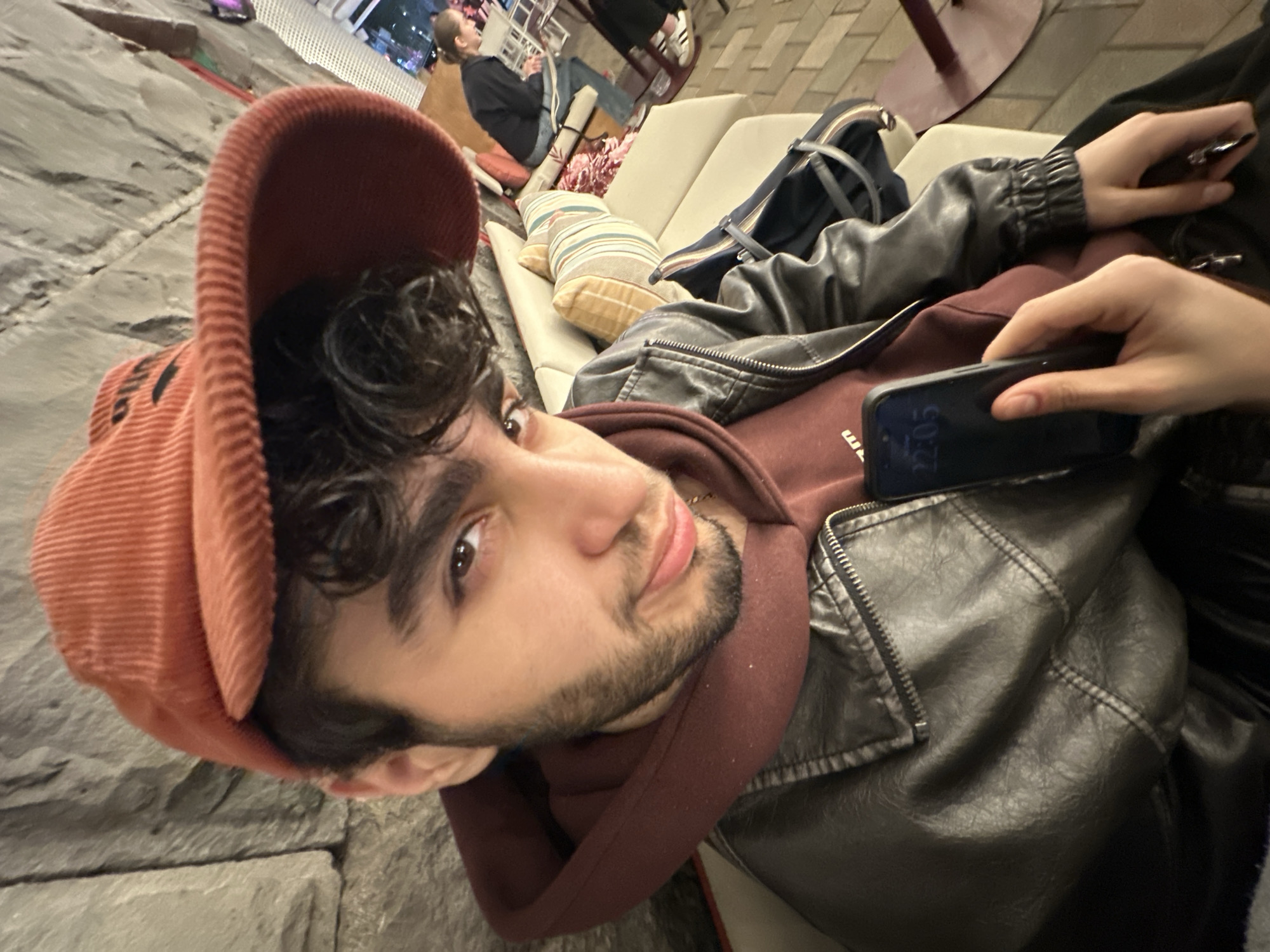 A young man with dark hair and a beard, wearing a burgundy hoodie, a black leather jacket, and a reddish-orange corduroy cap, is lying on a light-colored sofa. He is looking at the camera. Another person's hand is holding a black smartphone near him. In the background, people are sitting on a sofa with patterned cushions, and a bag is placed nearby. The picture was taken in Yerevan, Armenia.
