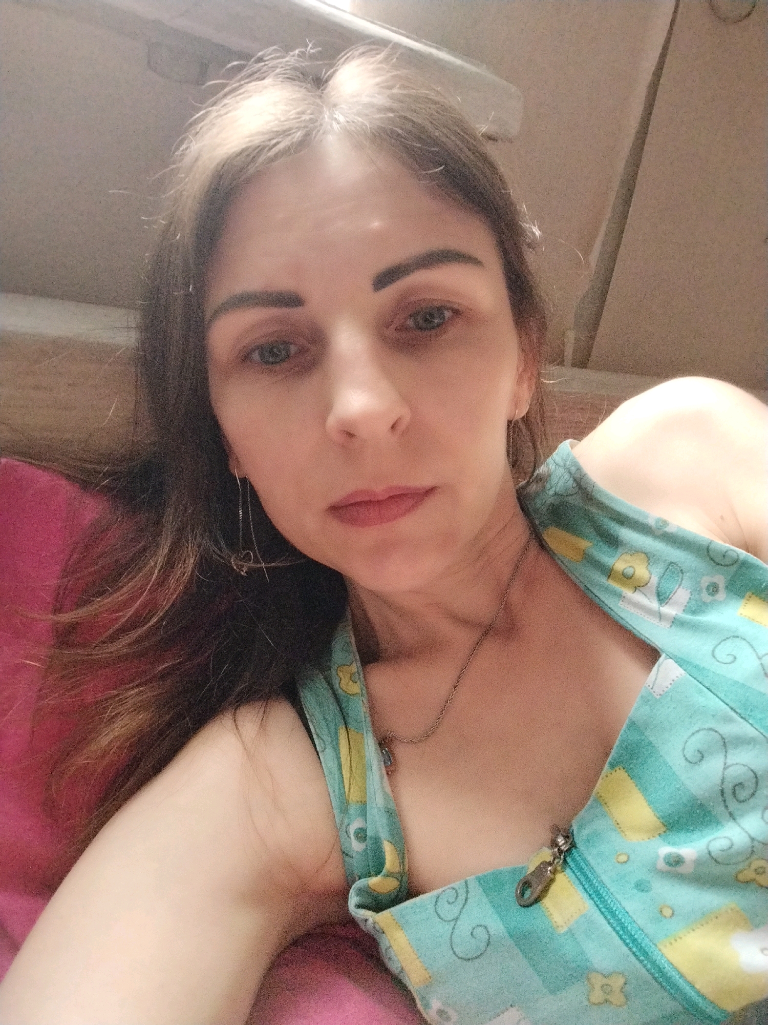 A woman with brown hair is lying down, propped up on her left arm, facing the camera. She is wearing a light blue top with yellow and white floral patterns and a zipper. She has a delicate necklace on. Her left ear has a dangling earring. The background is blurred, but appears to be an indoor setting, possibly a room. There is a pink pillow or cushion under her left arm. There is no information in the image or metadata to indicate the location is Vinnytsia, Ukraine.