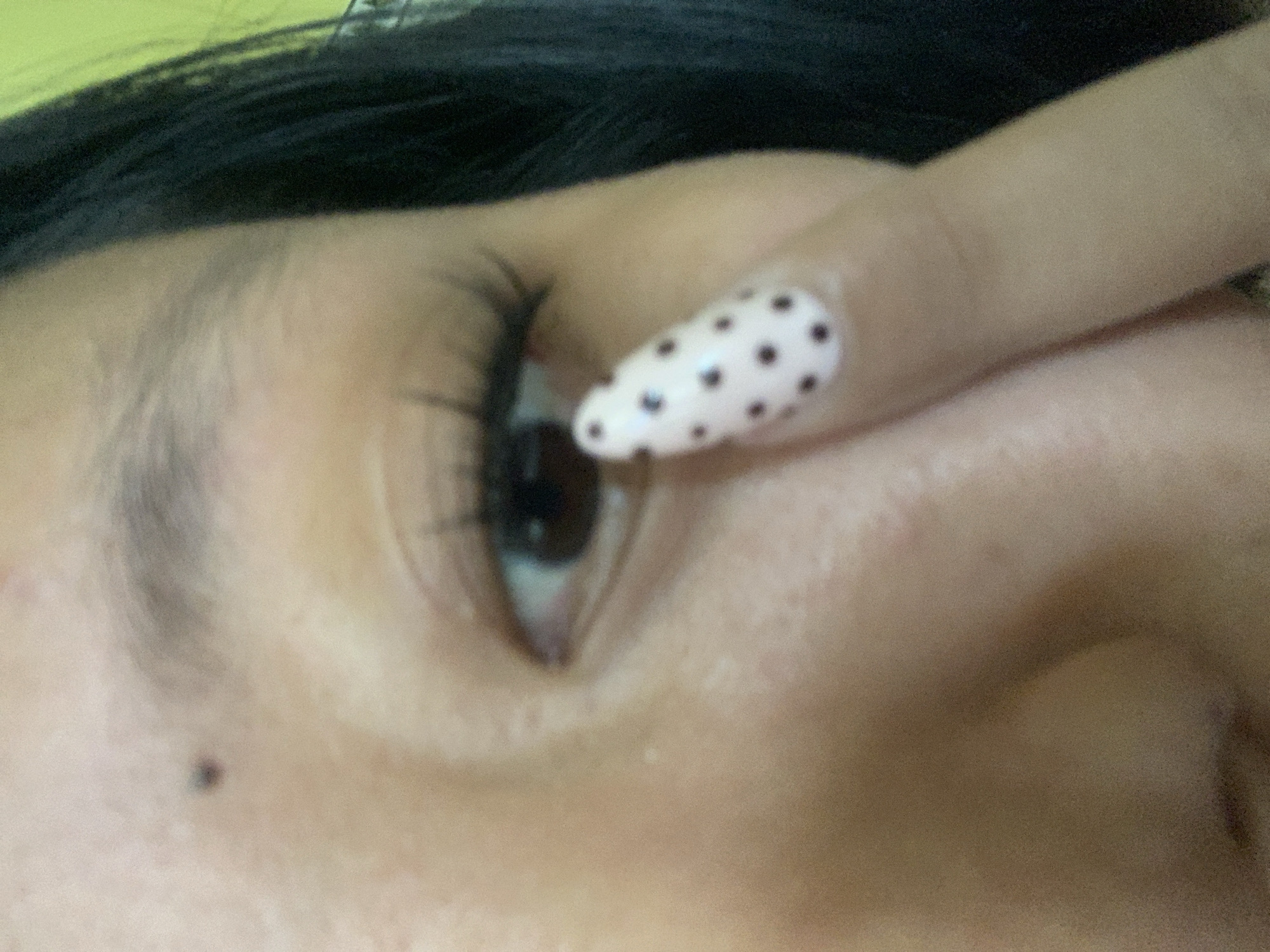 The image is a close-up of a person's eye and cheek, with a finger partially obscuring the eye. The person has dark hair and appears to be wearing false eyelashes. Their fingernail is painted with a light pink polish and decorated with dark polka dots. The background is a blurry green. There is no indication of the city of Jajag, Indonesia in the image.