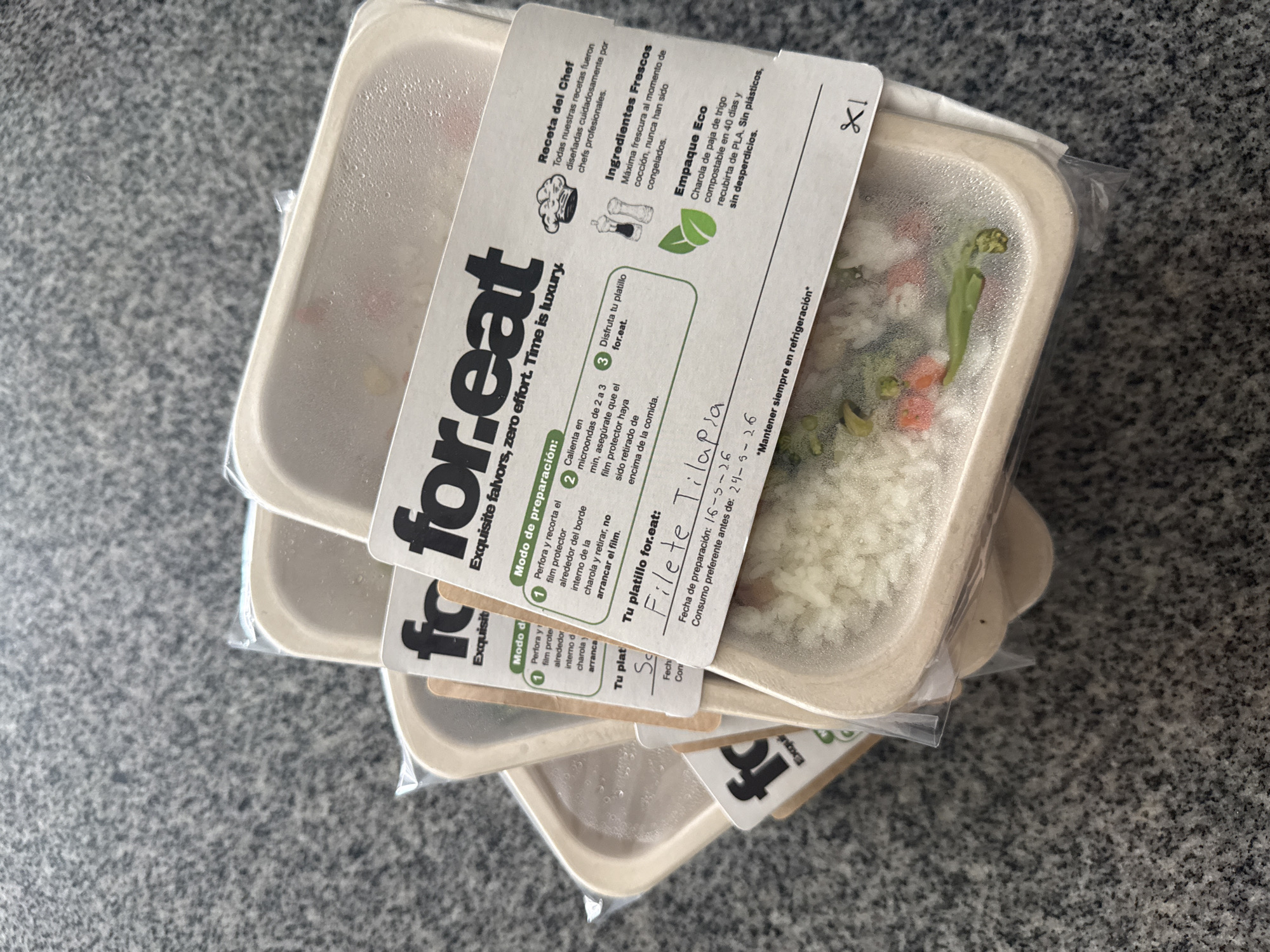 The image shows a stack of three ready-to-eat meal containers on a textured gray surface. The containers are made of a beige, possibly compostable material, and are sealed with clear plastic film. Each container has a white label wrapped around it, featuring the brand name "for.eat" in a modern sans-serif font. The labels also have information in Spanish, including cooking instructions, ingredients, and best-by dates. The top container's label indicates the meal is "Filete Tilapia" (Tilapia Fillet) with a preparation date of May 16, 2026, and a best-by date of May 29, 2026. Through the plastic film, one can see rice, vegetables, and possibly fish inside the container. The text on the label also states that the containers are eco-friendly and compostable and that they should be refrigerated.