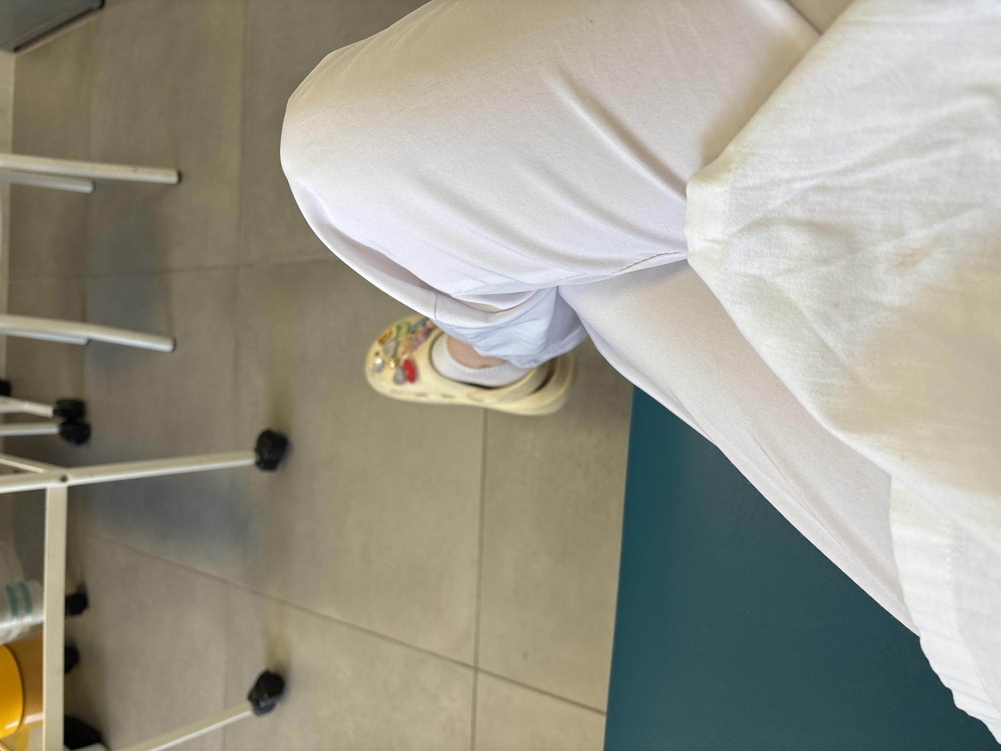 The image provides a top-down view of a person's lower body and surrounding environment, likely indoors. The subject is wearing white, possibly medical, trousers or scrubs, which appear slightly wrinkled. One foot is visible, clad in a light-colored (cream or pale yellow) closed-toe sandal or clog adorned with multiple colorful, decorative charms on its upper surface. A white sock is visible inside the sandal.
The floor beneath is composed of light beige or grey square tiles with darker grout lines, suggesting a clean, institutional setting. To the left side of the frame, part of a white metal structure, possibly a medical cart or stand, is visible, featuring horizontal bars and black plastic feet or wheels. Underneath this structure, a yellow cylindrical container and some white items are partially obscured. On the far right, a solid, dark teal or green surface, potentially a wall or large piece of furniture, provides a contrasting background element.
The overall environment, with its tiled floor, white attire, and utilitarian metal furniture, strongly implies a medical facility, such as a clinic or hospital. The person appears to be standing or seated in this space. No specific activity is taking place beyond their presence. There is no visible text, and the image does not provide cues regarding the time of day, weather, or the person's emotions. The location context is Saint Petersburg, Russia.