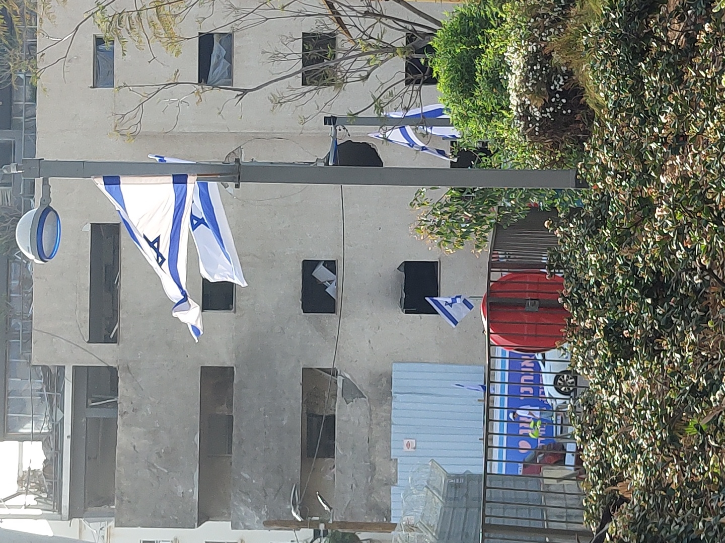 The image, taken in Tel-Aviv, Israel, displays a street scene featuring multiple Israeli flags against the backdrop of a building. Several white and blue Israeli flags, adorned with the Star of David, hang prominently from a grey street pole in the foreground. Smaller Israeli flags are also visible as decorations further down the street.
In the background, a light-colored, multi-story building appears to be under construction or renovation, characterized by its exposed concrete facade and numerous empty or unfinished window frames. Dense green foliage, including bushes and trees, frames the right side and top left of the image.
To the right, behind a black metal fence, a large red cylindrical object, likely a fire hose reel, is visible. Behind this fence, blue construction panels and a partially obscured white vehicle are present. A blue banner with white Hebrew text is also visible, reading "כביסה" (Laundry) and "ניקוי יבש" (Dry Cleaning), indicating a laundry and dry cleaning business.
The scene is illuminated by bright daylight, suggesting a clear, sunny day. The presence of numerous Israeli flags points to a display of national pride, possibly related to a national holiday or celebration, set within an active urban environment.