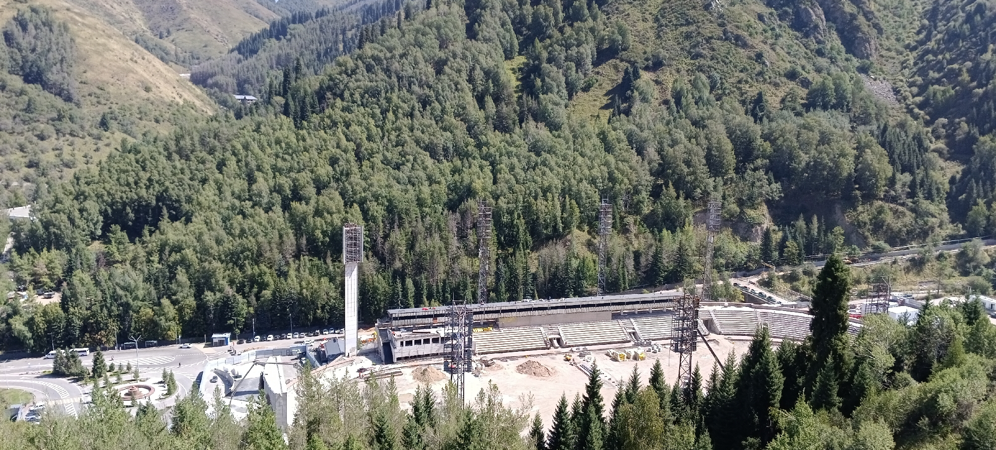 The image captures a wide shot of the Medeu Skating Rink in Almaty, Kazakhstan, nestled within a lush, tree-covered mountain valley. The high-altitude outdoor rink, famous for its speed skating track, is visible in the mid-ground, with tiered seating areas partially covered by scaffolding, indicating ongoing renovations or construction. Tall, industrial lighting towers stand sentinel around the rink, their metal structures stark against the green foliage and blue sky.
In the foreground, the natural environment dominates, with dense evergreen trees framing the scene and a winding road with a roundabout visible in the lower left, suggesting the approach to the sports complex. Cars and buses are parked along the road and within the complex's perimeter. The surrounding mountains are covered in a dense canopy of trees, with patches of dry, grassy slopes visible on the steeper inclines. The overall impression is of a significant sporting venue integrated into a dramatic natural landscape.