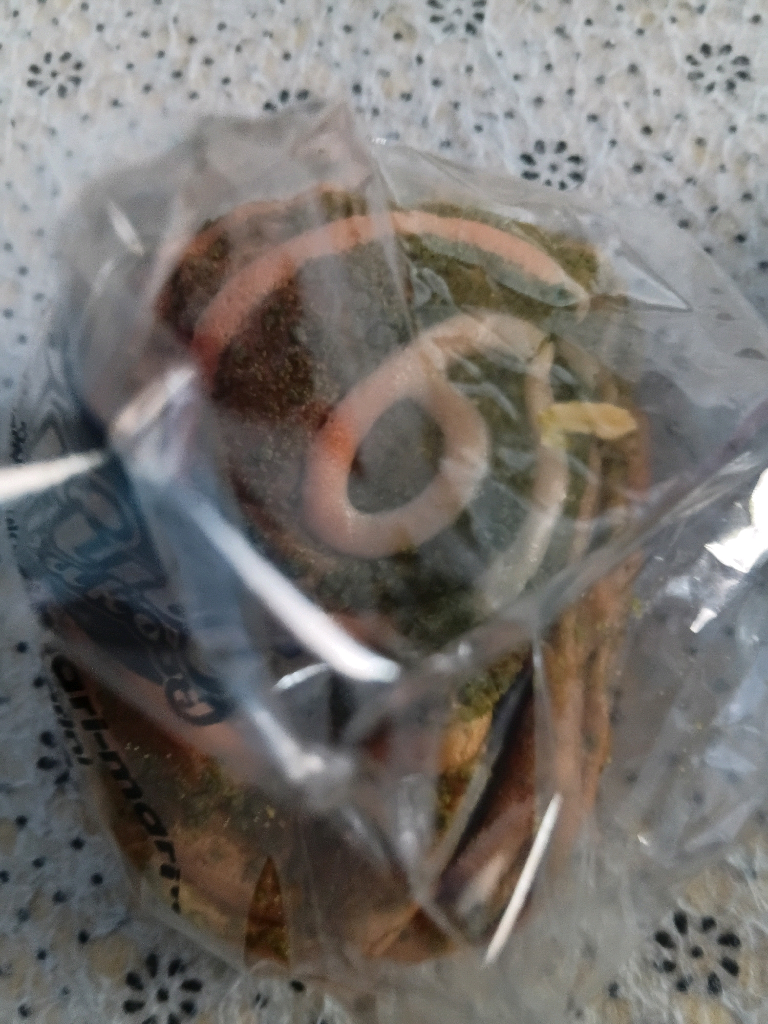 A pastry is displayed in a clear plastic wrapper, resting on a patterned tablecloth. The pastry, which appears to be a spiral-shaped roll, is covered in green specks and has a pale pink swirl on top. Text is visible on the plastic wrapper, partially obscured. The background is a light-colored tablecloth with a repeating floral or starburst pattern of black dots. The scene is located in Tanggul Wetan, Indonesia.