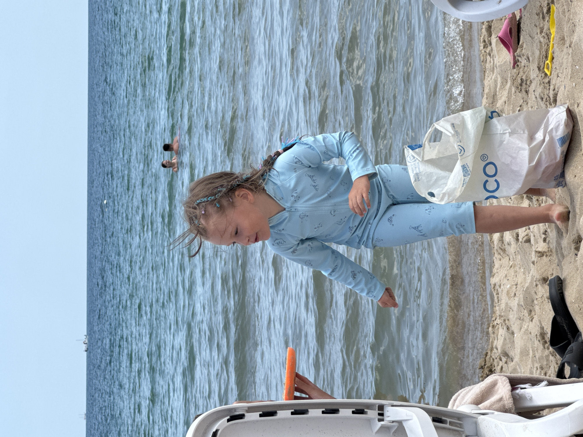 A young girl with braided hair stands on a sandy beach, wearing a light blue zip-up rash guard and matching bottoms decorated with a pattern of animals. She is looking to her left with her mouth slightly open. To her left, a person's hand is visible holding an orange frisbee. Beside the girl, a large white plastic bag with blue lettering is partially filled. In the background, the blue sea stretches to the horizon, with two small boats visible in the distance and two people swimming. The beach appears to be in a coastal area, likely a resort town, possibly Nesebar, Bulgaria, given the presence of people enjoying a beach environment and the general aesthetic.