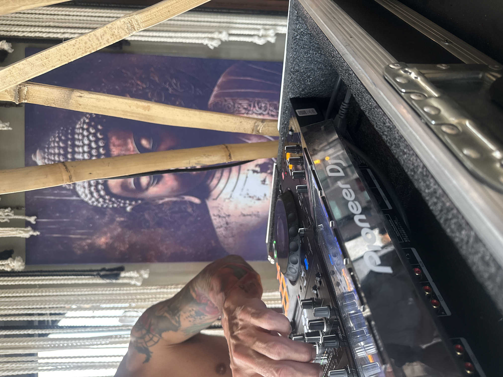 The image shows a person operating a Pioneer DJ mixer, housed in a protective case with metal trim. The person's hands are visible, and they are tattooed. Behind the mixer, there is a tapestry or poster featuring a Buddha image. Bamboo stalks are positioned in front of the tapestry. The environment is decorated with macrame hangings. The image includes Japanese writing on the back of the mixer. The scene is taken in Mexico City, Mexico.