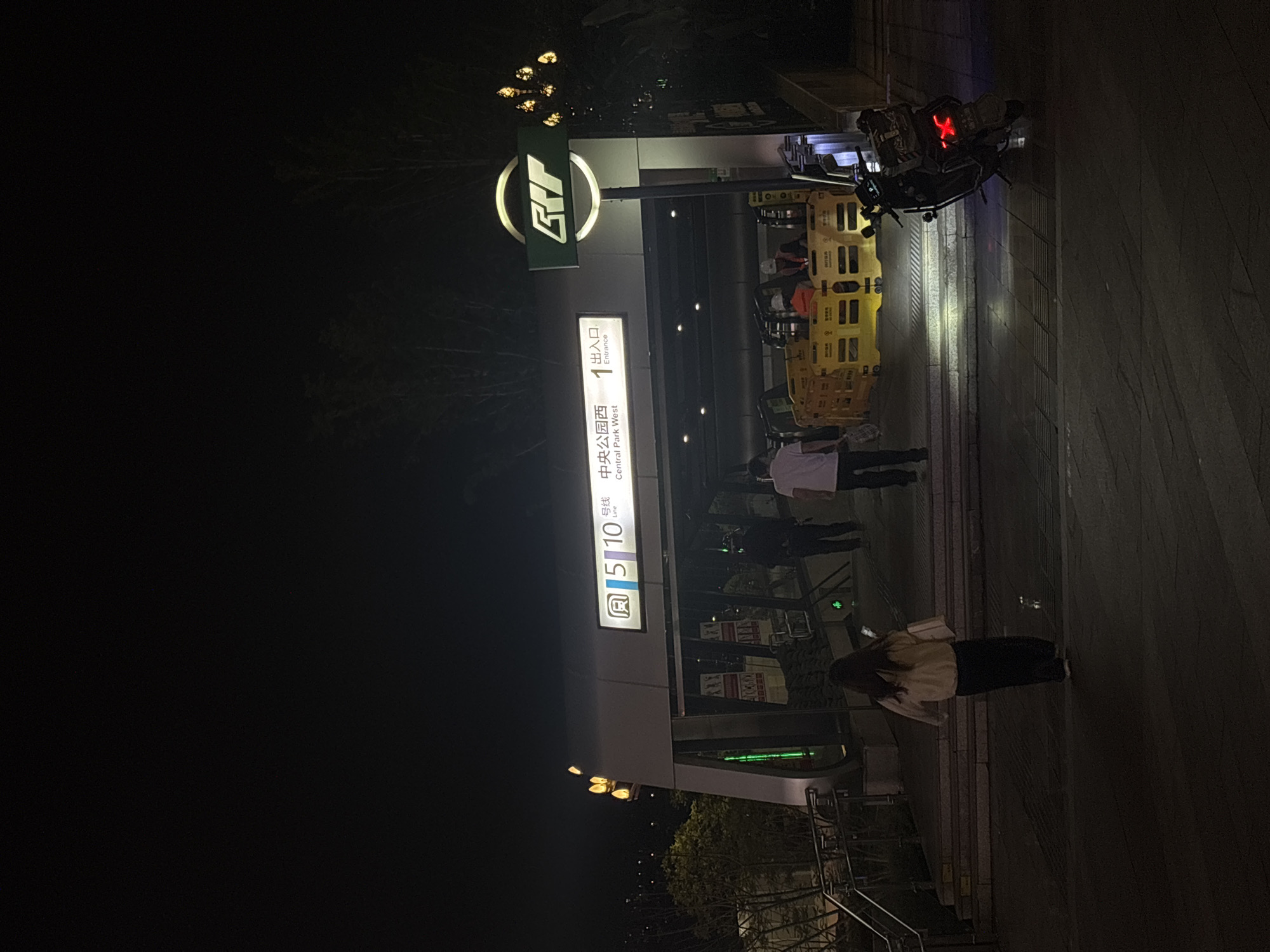 At night, the entrance to the Central Park West station (Exit 1) of the Chongqing Rail Transit Line 5 in Liangjiang, China, is visible. People walk along the sidewalk by the entrance. A luggage cart sits by the entrance.