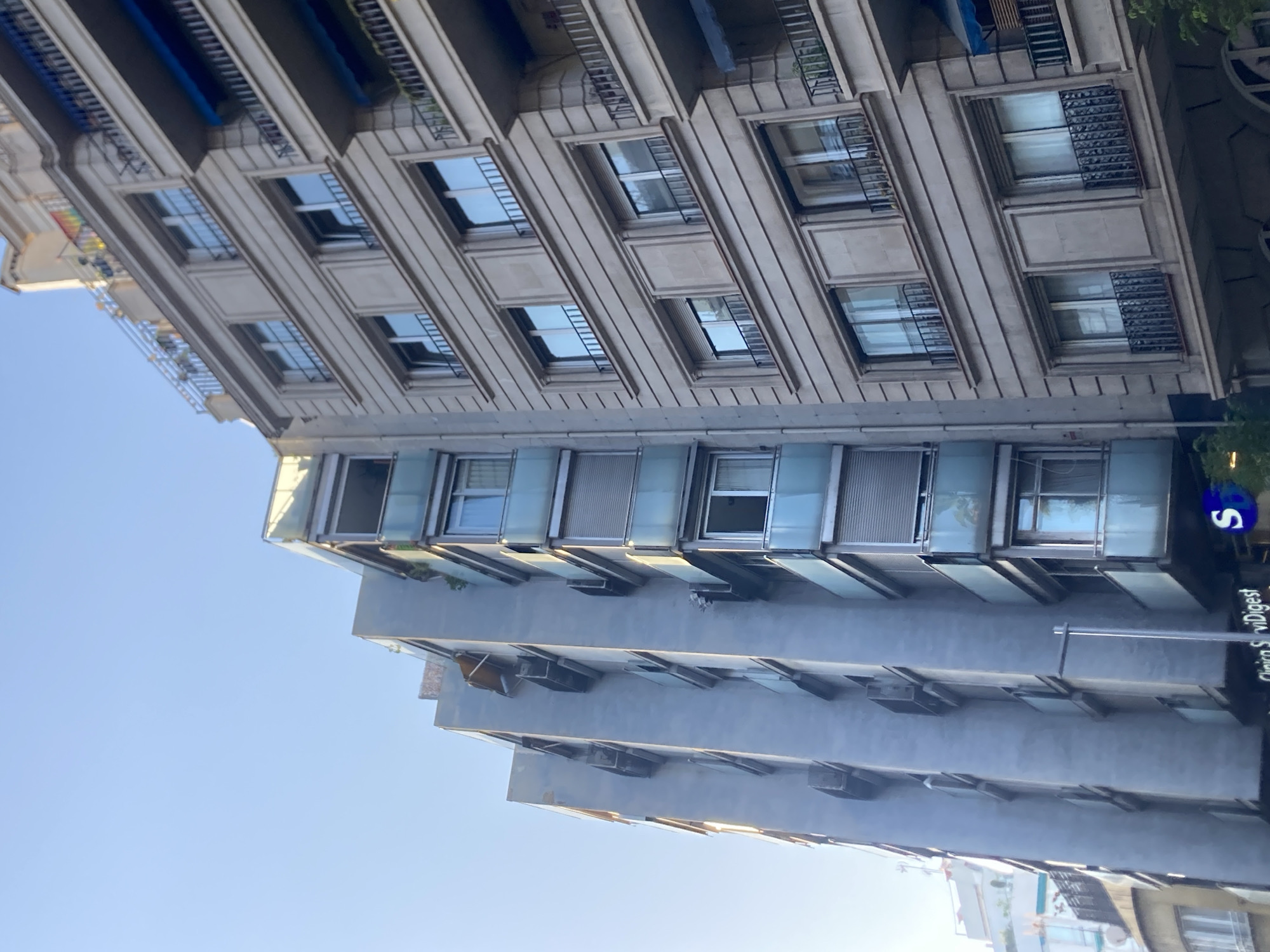 This is a vertical, wide-angle photograph of buildings in Barcelona, Spain. The image captures the architecture of several stories of two connected buildings. The building on the left appears to be older, with cream-colored stone and numerous windows with decorative grates. The building on the right is more modern, with a gray facade and a series of glass-enclosed balconies or rooms that project outward. The sky is clear and blue, suggesting a daytime shot, likely in the morning or afternoon due to the lighting.
The scene is static and there are no people visible. The focus is on the urban environment and the contrast between the architectural styles of the two buildings. Notable details include the reflective surfaces of the modern building's windows and the patterned grates on the older building's windows. A blue circular sign with a white "S" is visible on the right side of the modern building, possibly indicating a business or establishment. Below the sign, some text is partially visible: "Clinica S" and "Digest". The overall impression is one of a well-lit, clear day in a European city.