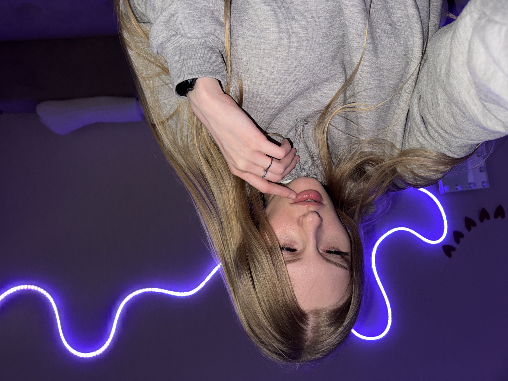 This media file captures a young person with long, blonde hair, posed close to the camera. The image is taken from a low angle, with the person's head and upper body filling most of the frame. They are wearing a grey hooded sweatshirt. One hand is brought up to their lips, with a ring visible on one finger. Their eyes are closed, and their expression could be interpreted as thoughtful or sensual.
The scene is illuminated by a wavy strand of purple neon light, which curves across the lower part of the image. In the background, a dark purple wall is visible, with some decorative heart-shaped elements on the right side. A white, fluffy object, possibly a pillow or blanket, is also present in the upper left corner. The overall mood is intimate and somewhat mysterious, enhanced by the low lighting and the glow of the neon. The time of day is likely evening or night due to the artificial lighting. There is no visible text in the image.