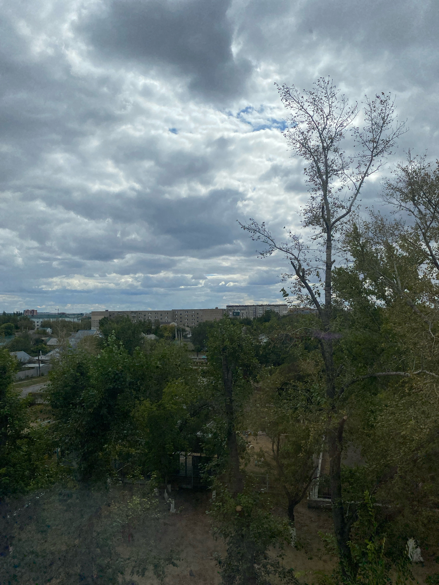 The image depicts an overcast sky over a residential area in Kostanay, Kazakhstan. In the foreground, several trees are visible, some with sparse foliage, suggesting it might be autumn. Beyond the trees, a collection of buildings can be seen, including multi-story apartment blocks and smaller houses. The sky is filled with heavy, gray clouds, with a small patch of blue visible in the upper center. The overall impression is a somewhat muted, possibly cool-weather scene.