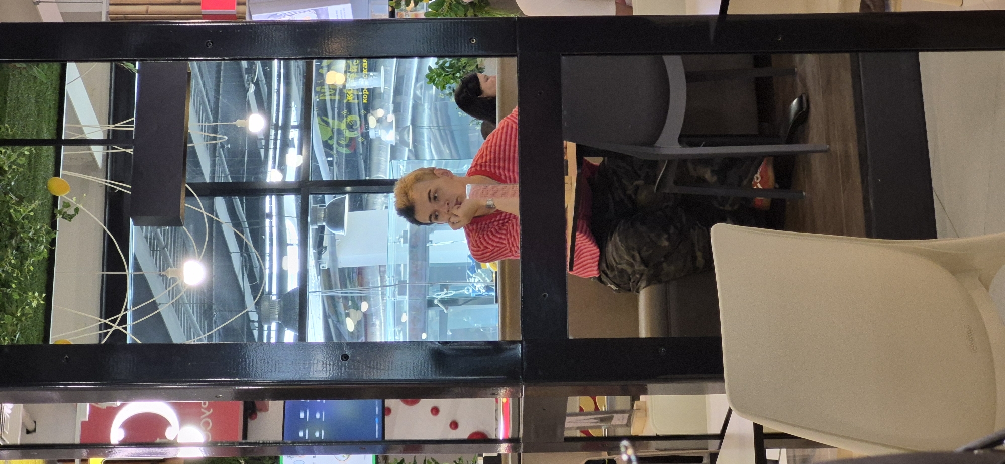 The image depicts an indoor scene, likely a cafe or restaurant in Vladivostok, Russia, as suggested by the Russian text visible on a sign reflected in the glass. The primary subject is a person with blonde hair, partially dyed black, wearing a red and white striped shirt and camouflage pants, seated at a table. They are resting their chin on their hand and looking off to the side. Another person with dark hair is visible in the background, also seated. The environment includes tables, chairs, and overhead lighting fixtures. The image is framed by black structural elements and reflections of the interior and ceiling are visible in the glass.