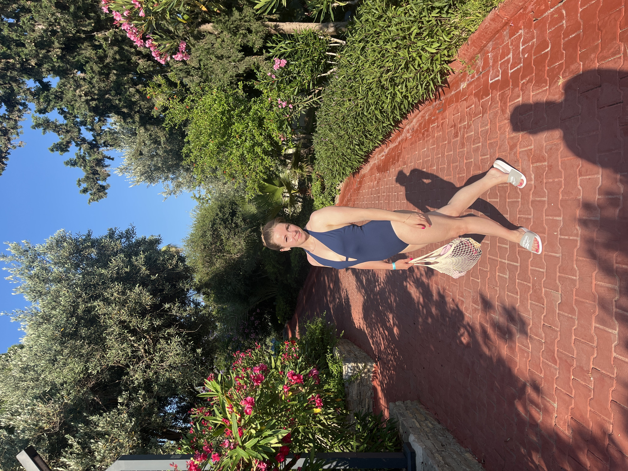 A woman stands on a red brick pathway lined with lush green bushes and flowering plants. She is wearing a navy blue one-piece swimsuit and holding a white mesh bag. The pathway curves into the distance, surrounded by trees and greenery under a clear blue sky. The scene appears to be a sunlit outdoor area, likely within a resort or park, given the landscaping. The location is identified as Alanya, Turkey.
