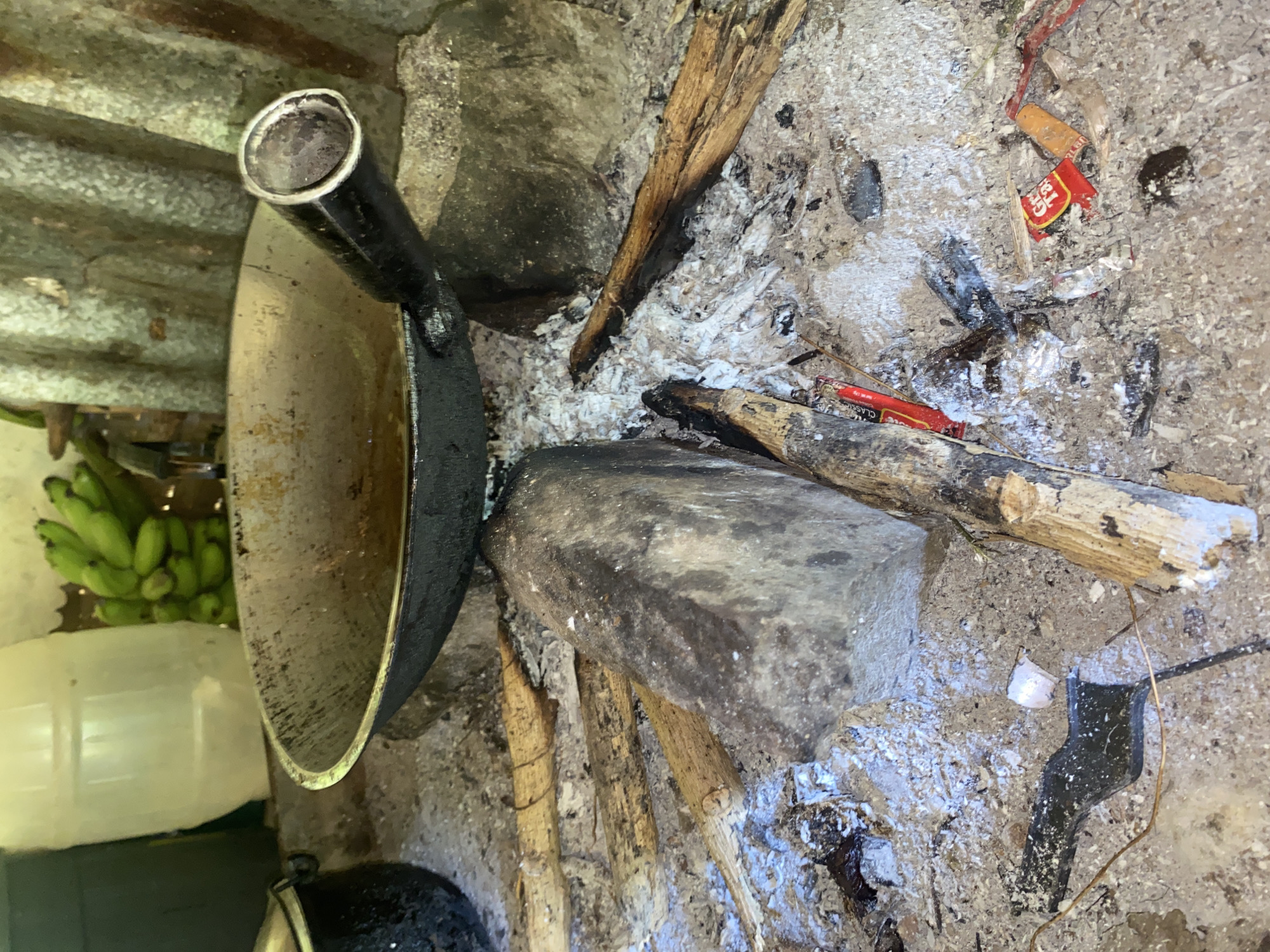 The image depicts a rustic outdoor cooking setup. A well-used wok sits over a bed of hot coals and ash, with several pieces of firewood scattered around. In the background, a bunch of green bananas hangs, and a large plastic container is visible. The scene suggests a cooking area, likely in a rural or less developed setting. The presence of ash, embers, and firewood indicates recent or ongoing cooking activity. The corrugated metal structure behind the wok suggests a shelter or wall. The overall impression is one of simple, functional food preparation. Based on the provided context, this scene is located in Davao City, Philippines.