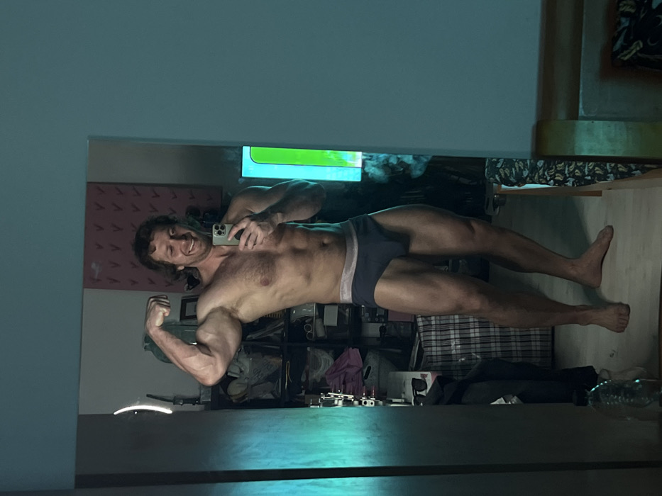 A male adult with medium-length brown hair is seen reflected in a mirror, taking a selfie with a smartphone held in his right hand. He is shirtless, wearing dark blue briefs with a light grey waistband, and is barefoot. He stands with his left arm flexed, showcasing his bicep, and is smiling broadly towards the camera.
The background visible in the mirror's reflection is an indoor, cluttered domestic space. To the subject's left, a shelving unit or open wardrobe contains various items, including folded clothing, what appears to be bags, and sunglasses. A large checkered storage bag or laundry basket is visible at the bottom left. Above the shelving unit, a reddish-pink rectangular object with a pattern of small "V" shapes is mounted on the wall. To the subject's right, a vertical glowing light fixture or screen displays a combination of bright green and light blue light. Further right, a portion of a bed with a patterned sheet is visible. The overall lighting is dim, with a prominent teal-cyan light source illuminating the left side of the scene.
The image provides no visual indicators such as landmarks, street signs, or unique architectural features to specify a precise location within the city of Saint Petersburg, Russia.