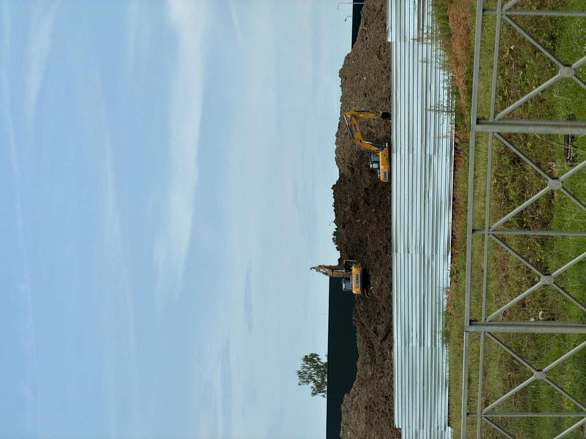A construction site in Bersenevka, Russia, features two yellow excavators amidst large piles of brown soil under a pale blue sky with scattered clouds. In the foreground, a metal fence with a diamond pattern is visible, with green grass and some vegetation growing behind it. A corrugated metal barrier runs horizontally across the middle ground, separating the foreground from the construction area. A dark fence is visible in the background, and a solitary tree with green foliage stands to the left of the excavators. The overall scene depicts ongoing construction or excavation work in an outdoor environment.