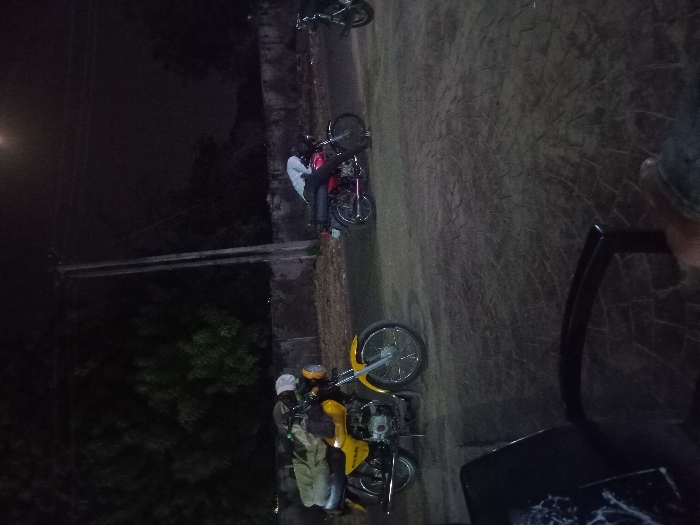 Nighttime scene featuring two motorcycles parked on a dirt path alongside a narrow stone wall. One motorcycle is yellow and black, the other red and black. People are visible near the motorcycles. Overhead electrical lines stretch across the sky. The overall lighting is dim, with a light source illuminating the upper left area. The perspective suggests the photo was taken from an elevated position.