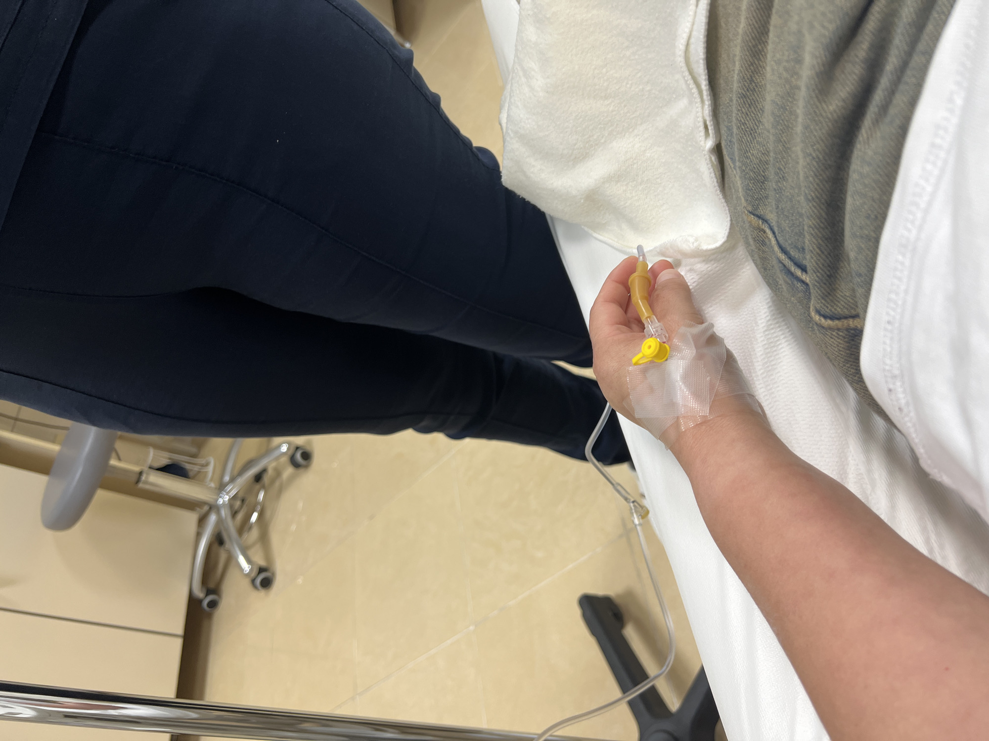 An overhead, high-angle shot depicts an indoor scene in what appears to be a medical facility in Podgorica, Montenegro. The central subject is a person's left forearm and hand, where an intravenous (IV) cannula, secured with clear tape and featuring a yellow cap, is inserted into the back of the hand. IV tubing extends from the cannula, indicating an ongoing or recently initiated medical infusion. This individual seems to be a patient, lying or seated, partially covered by a white sheet or blanket, with part of their lower body showing a greyish-brown denim fabric.
To the left, another person is seated, with their legs visible, clad in dark blue trousers. Below them, a chrome-plated, five-star rolling chair base with wheels is prominent on the light-colored tiled floor. Also on the floor are parts of a black equipment stand and a shiny metallic bar, possibly part of a bed frame or other medical apparatus. The environment appears clean and functional, consistent with a clinic or hospital room. No specific time of day is indicated, and there is no visible text.