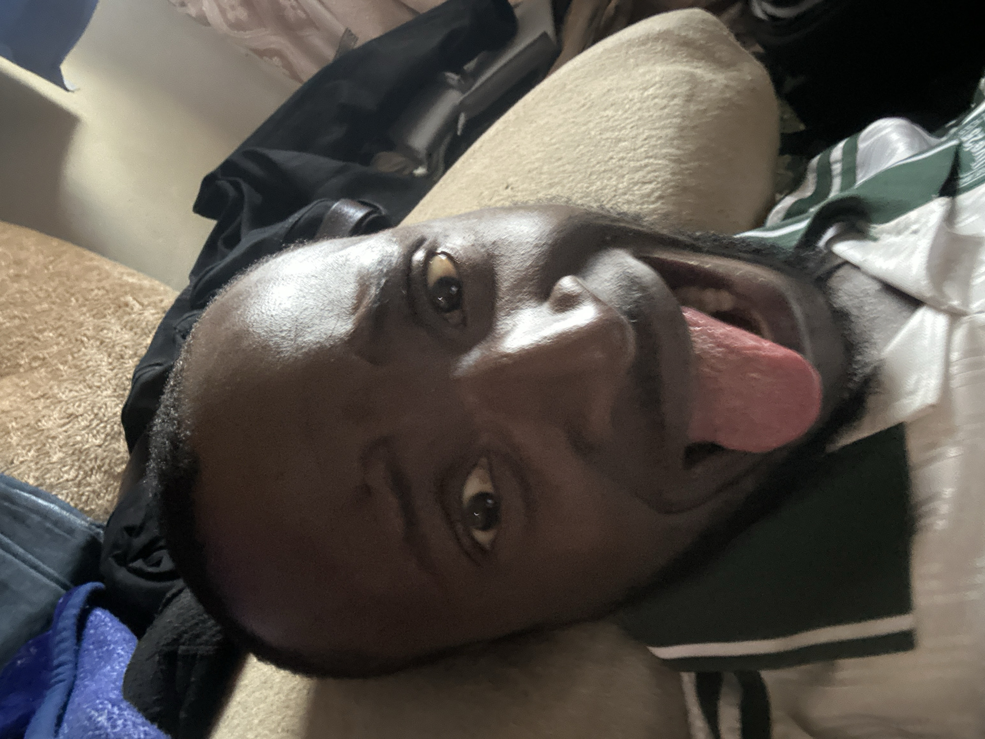A man with dark skin and a shaved head is lying down, looking directly at the camera with his tongue sticking out and eyes wide. He is wearing a white shirt with dark green and white stripes around the collar. The background is blurry, with what appears to be bedding or soft furnishings. The image is taken from a close-up, low-angle perspective. There is no discernible information within the image to indicate the location in Jos, Nigeria.