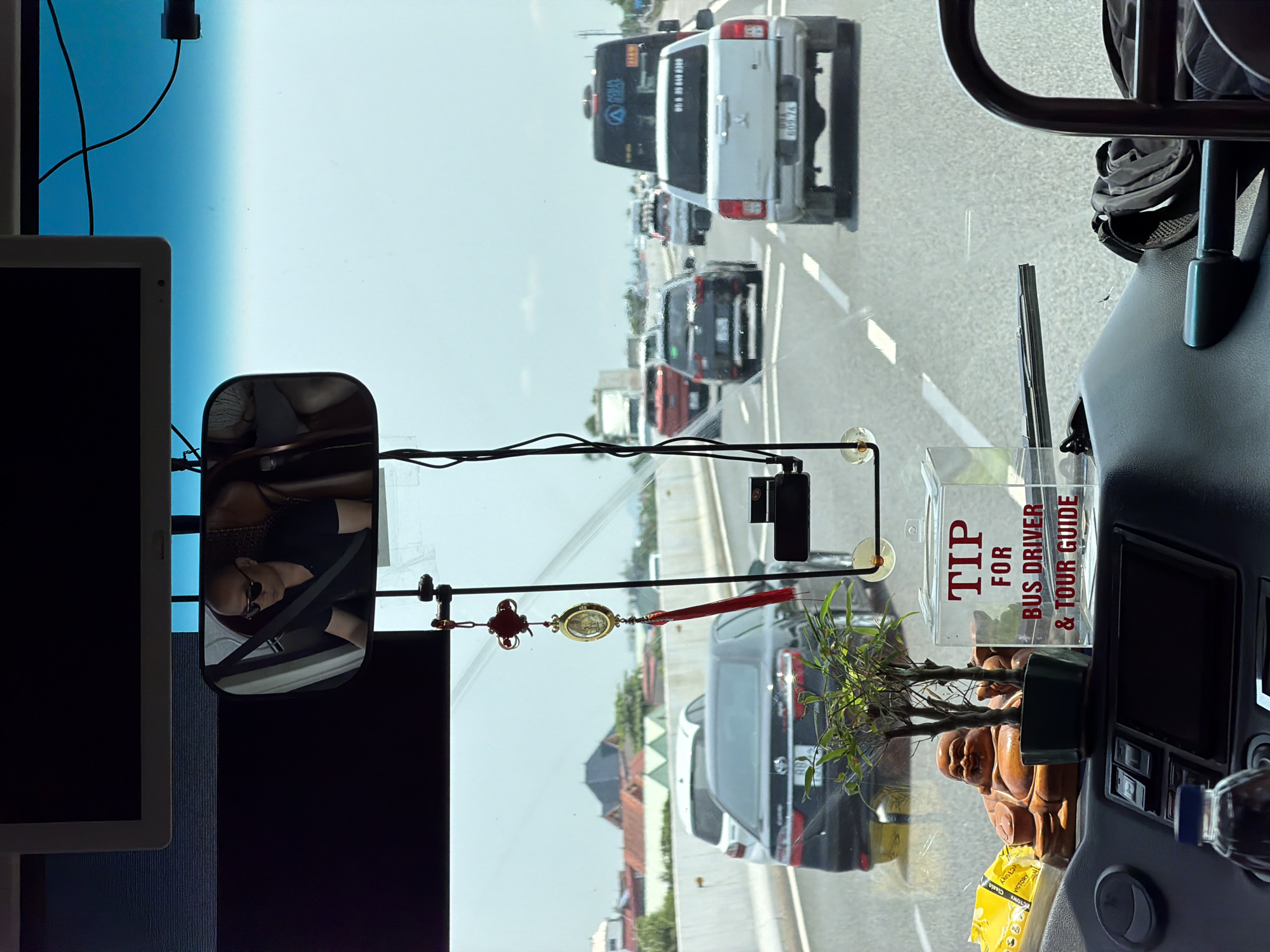 The image provides a view from inside a vehicle, likely a bus or large van, looking out onto a highway in Hải Phòng, Vietnam, under a clear daytime sky.
In the foreground, inside the vehicle, various objects adorn the dashboard and windshield. On the right, a dark control panel with buttons and a screen is visible. Next to it, a small potted plant, resembling bamboo, sits beside a Laughing Buddha figurine. A clear plastic box with prominent red text reads "TIP FOR BUS DRIVER & TOUR GUIDE," suggesting the vehicle is used for tours or public transport. A yellow snack package labeled "VICTORY Classic" is also on the dashboard, along with some pens in a container.
An ornate red and gold charm or tassel hangs from the windshield, near a small black dashcam. The rearview mirror reflects a person, likely the driver or a passenger, wearing sunglasses and a seatbelt. To the left, a portion of a vertical monitor screen is visible. The upper part of the windshield displays a gradient of light blue and white.
Outside the vehicle, a multi-lane highway is filled with moderate traffic. Several cars, a white SUV or truck, and a large black bus are visible ahead. The black bus clearly displays "HÀ MINH THÀNH" and a company logo on its side. The scene depicts a typical travel setting on a sunny day.