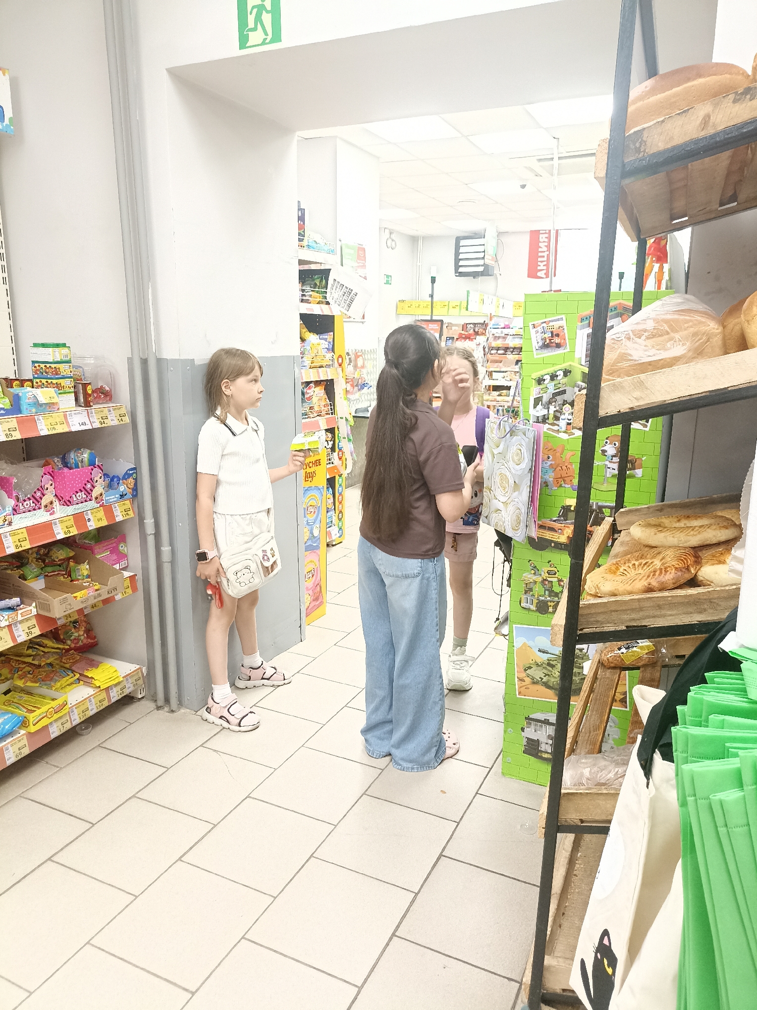 In a retail environment resembling a small grocery store or a section of a larger supermarket in Tyumen, Russia, three individuals are observed. A young girl, appearing to be between 8 and 10 years old, stands on the left side of the frame, facing right. She is wearing a white short-sleeved top, a light-colored skirt, white socks, and pink sandals. A small crossbody bag with a teddy bear design is attached to her waist. She holds a small object in her right hand, possibly a toy or a candy.
To her right, a second girl, approximately 12 to 14 years old, is positioned closer to the center. She is wearing a pink t-shirt, light-colored shorts, and white sneakers. Her hair is pulled back in a ponytail, and she is looking towards the viewer's left.
Partially obscured behind the second girl, and to her right, is a third individual, likely an adult woman, wearing a brown t-shirt and blue jeans. She is facing towards the second girl, and her left arm is raised, with her hand near the second girl's face. Their interaction appears casual and possibly playful.
The environment is characterized by shelves stocked with various products, including packaged goods, candies, and baked items. On the far right, a shelf displays bread and other baked goods. The flooring is tiled with a light-colored grid pattern. An emergency exit sign with a green running figure is visible in the upper left quadrant of the image. A red sign with white Cyrillic text, likely an advertisement or sale notice, is visible further in the background. The overall lighting suggests an indoor commercial setting.