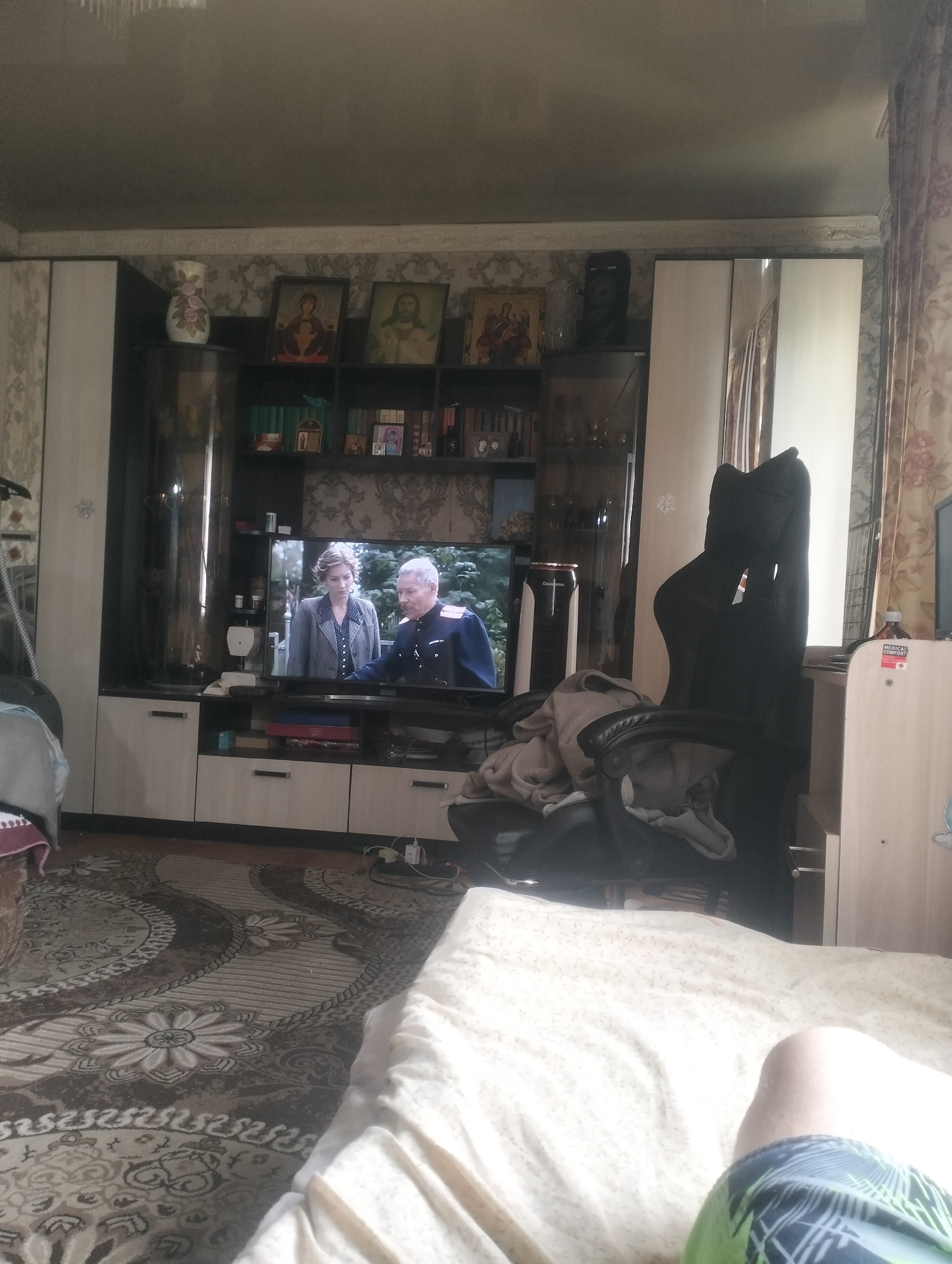 The image depicts a living room scene. A large television screen displays a program featuring two individuals, a woman in a grey jacket and a man in a military uniform. The television is part of a dark wood entertainment unit, below which are several drawers and cabinets. Above the television, on shelves, are various decorative items, including framed religious icons and what appears to be a vase. To the right of the entertainment unit is a tall black office chair with clothing draped over it. Further to the right, a tall, narrow white cabinet with a mirrored door is visible. The room has patterned wallpaper and a patterned rug on the floor. In the foreground, a person's legs, clad in shorts with a green and blue pattern, are visible resting on a white comforter, suggesting the photo was taken from a bed. The general impression is that of a lived-in, domestic interior. The location is specified as Almaty, Kazakhstan.