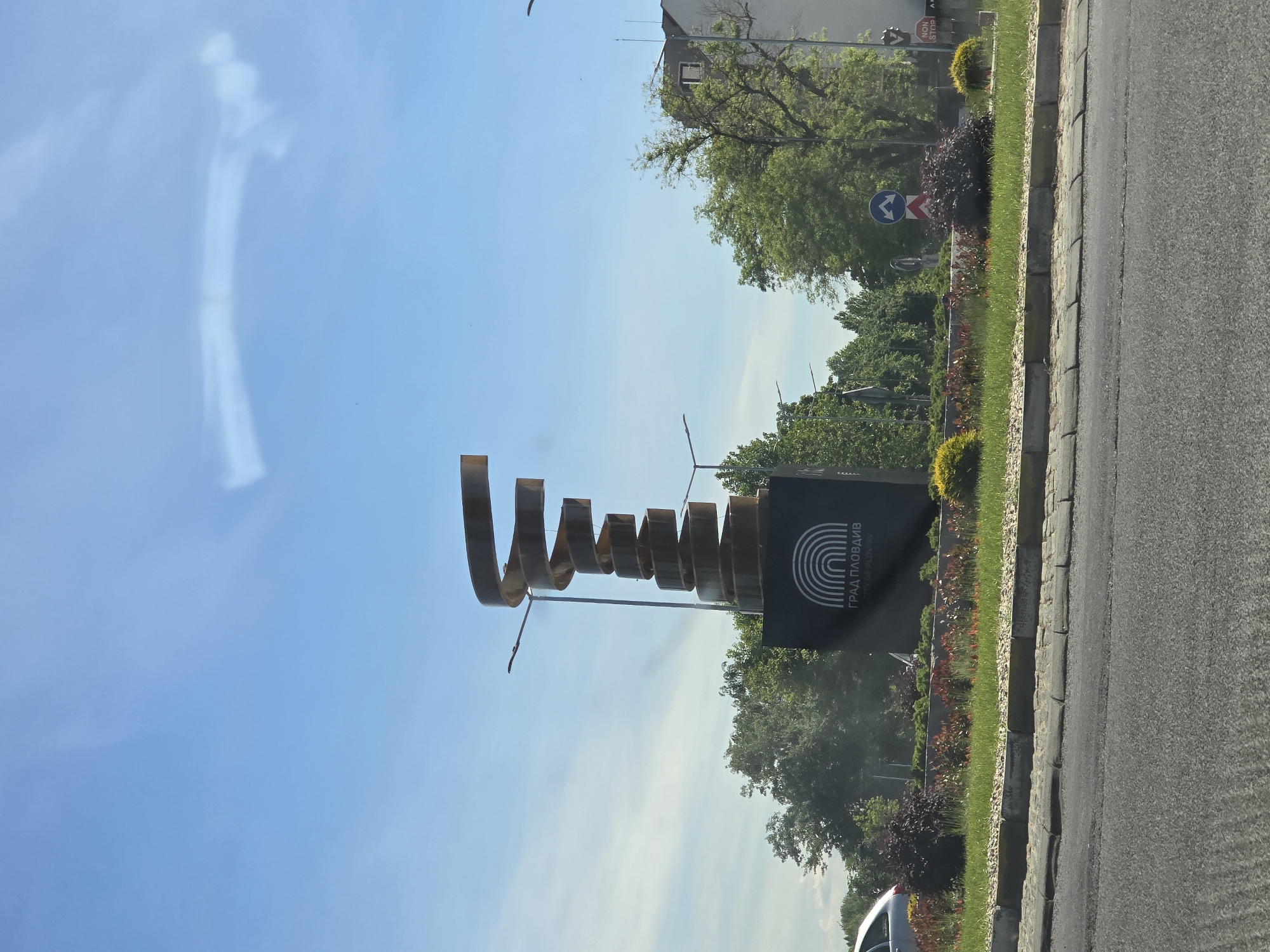 The image shows a city welcome sign situated on a median. The sign consists of a black rectangular base with an abstract metal sculpture extending upwards. The black base has a stylized logo of concentric arcs, with the text "ГРАД ПЛОВДИВ". The background includes trees, green foliage, and a blue sky with some clouds. The location is in the city of Plovdiv, Bulgaria. A road is adjacent to the median.