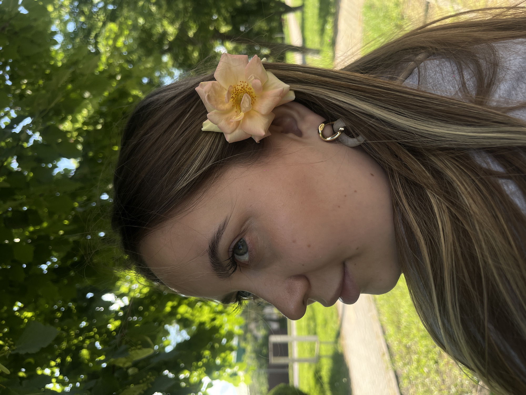 A young female subject is depicted in profile, facing the viewer's left, in an outdoor setting within the city of Novopokrovskaya, Russia. Her long, straight to slightly wavy, medium brown hair features prominent blonde highlights, extending over her right shoulder. A single, light peach-toned rose, displaying yellow stamens, is positioned behind her right ear. She wears a two-part earring on her right ear, comprising a small gold-colored hoop connected to a larger, opaque, light grey, crescent-shaped pendant. Her left eye, characterized by a light blue or grey iris and notably long eyelashes, is visible and directed slightly upwards and to the left. Her nose has a straight bridge and slightly upturned tip, and her lips are slightly parted. The visible portion of her attire is a light grey top. The background consists of a dense canopy of green leaves above, through which dappled sunlight creates bright, out-of-focus light circles. Below, green grass is visible, along with a blurred paved path and a faint, light-colored rectangular structure in the mid-ground distance.