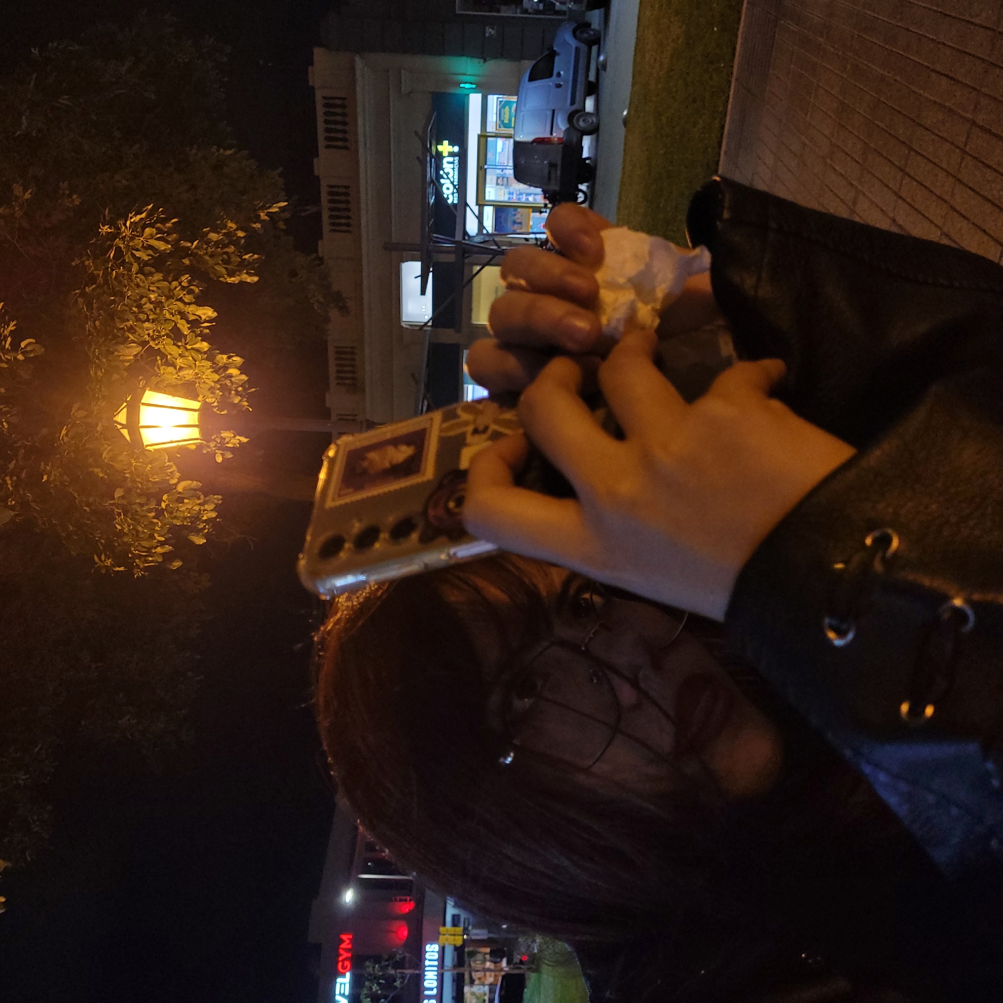 A person is taking a photo with a smartphone at night in San Fernando del Valle de Catamarca, Argentina. The person is wearing glasses, and a black jacket with metal rings. The phone has a decorative case. The person is holding a white tissue in their hand. In the background, there's a building with a pharmacy (Farmacias Colon) and a light blue vehicle parked nearby. A street light illuminates the scene and a tree. A gym (VEL GYM) is visible in the background.