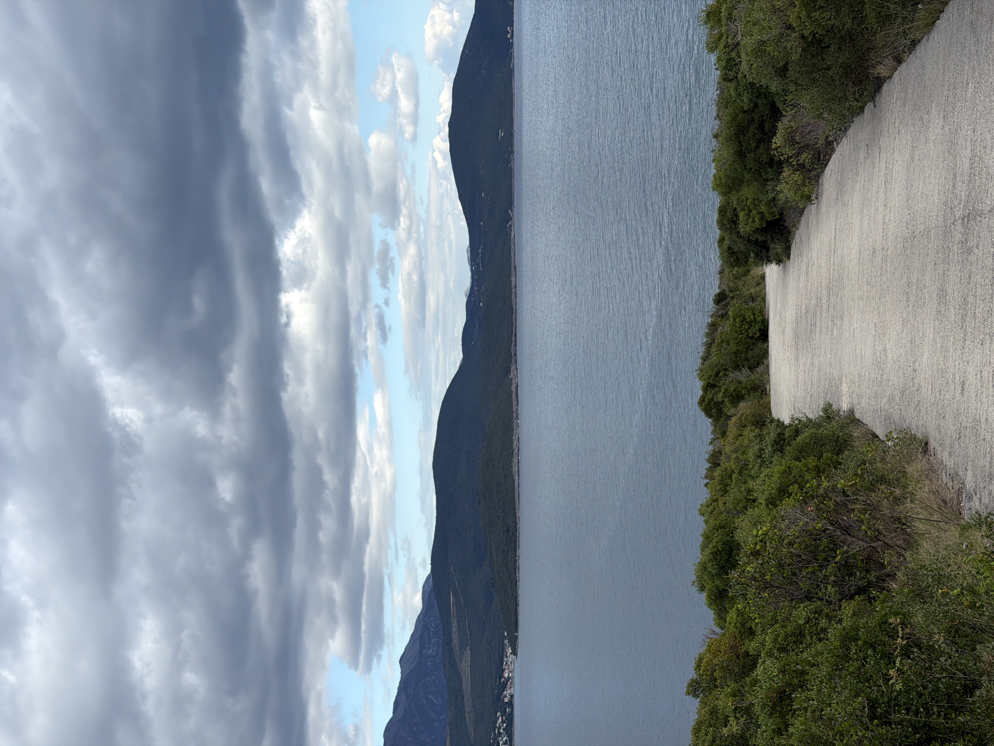 The image depicts a coastal scene, potentially along the Adriatic Sea. A paved road is seen on the right side of the image, bordered by lush green vegetation. To the left of the vegetation, the image shows the sea with mountainous land visible across the water. The sky is filled with clouds, some dark and ominous, while others are lighter with patches of blue sky showing through. The image is in Radovići, Montenegro.