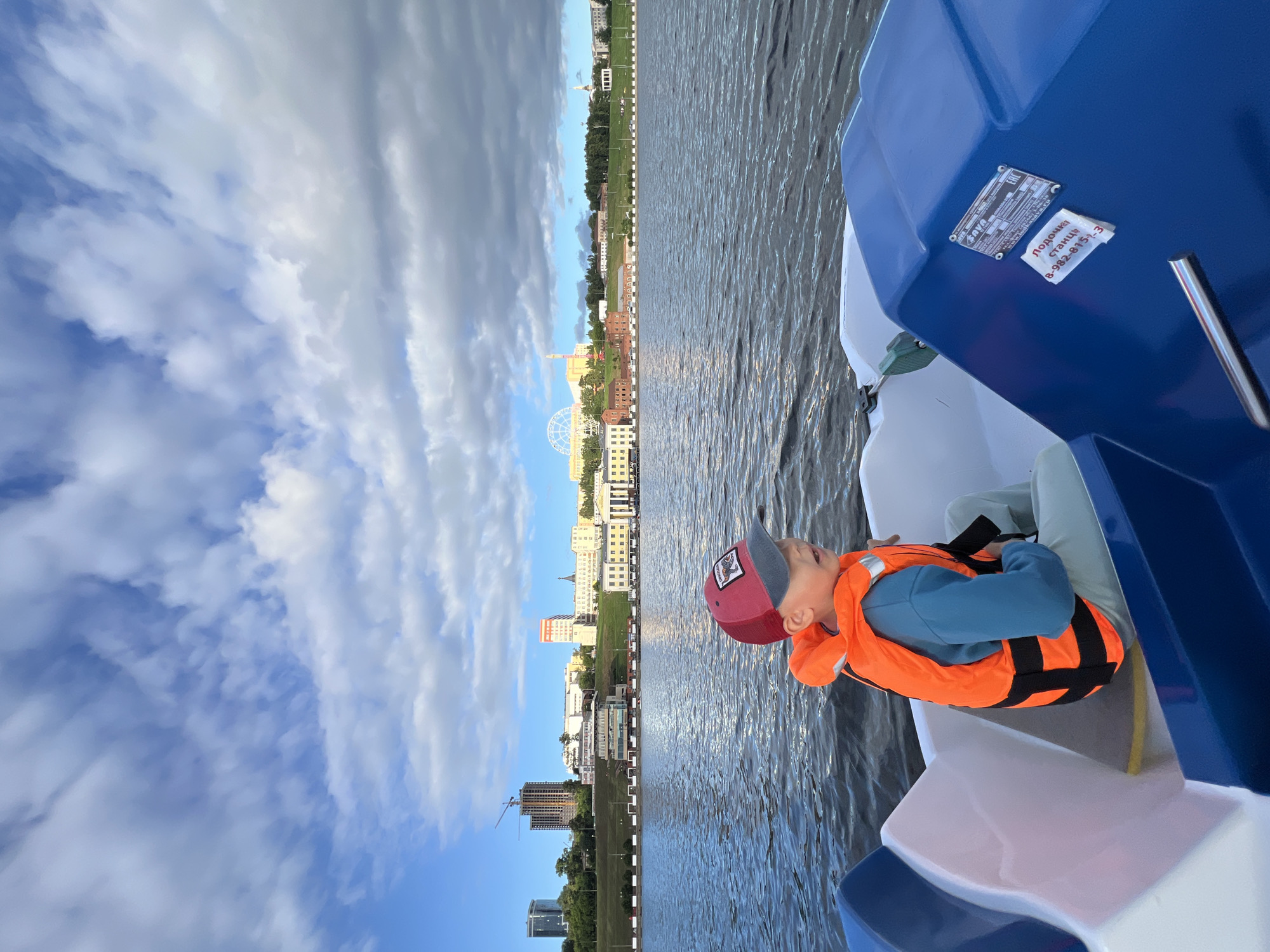 The image captures a young boy wearing a red and grey baseball cap and an orange life vest as he sits in a blue and white pedal boat. He is looking upwards, seemingly enjoying the view. The boat is on a body of water, with a city skyline visible in the background. The skyline features various buildings, a Ferris wheel, and a construction crane, suggesting an urban environment. The sky is partly cloudy with a mix of blue sky and white clouds, indicating daytime, likely late afternoon or early evening given the soft light. The water is rippled, suggesting a gentle breeze. On the blue part of the boat, there is a metal plate with some text and a small white sticker with Cyrillic text and a phone number. The text on the sticker appears to read "Полочка станц" followed by a phone number. The overall mood is relaxed and pleasant, with the child appearing engaged and happy in the outdoor setting of Izhevsk, Russia.