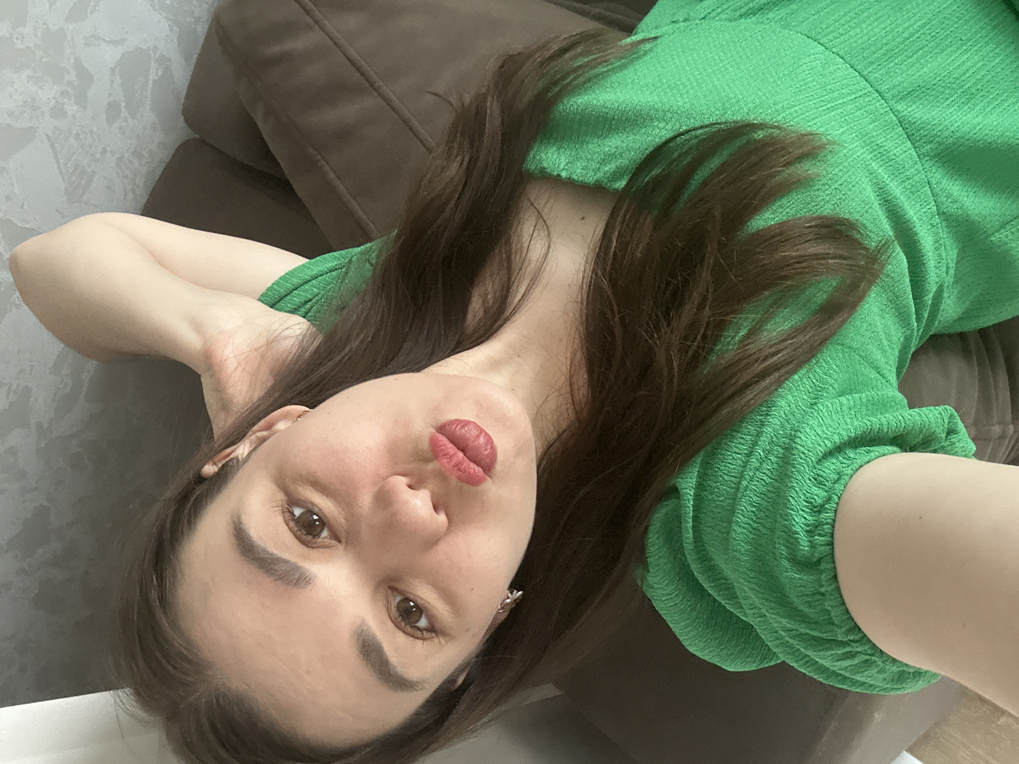A woman lies on a brown couch. She is wearing a green dress and has brown hair. The wall behind the couch is a light grey. The location in the city of Chaikovsky, Russia is not discernible from the image.