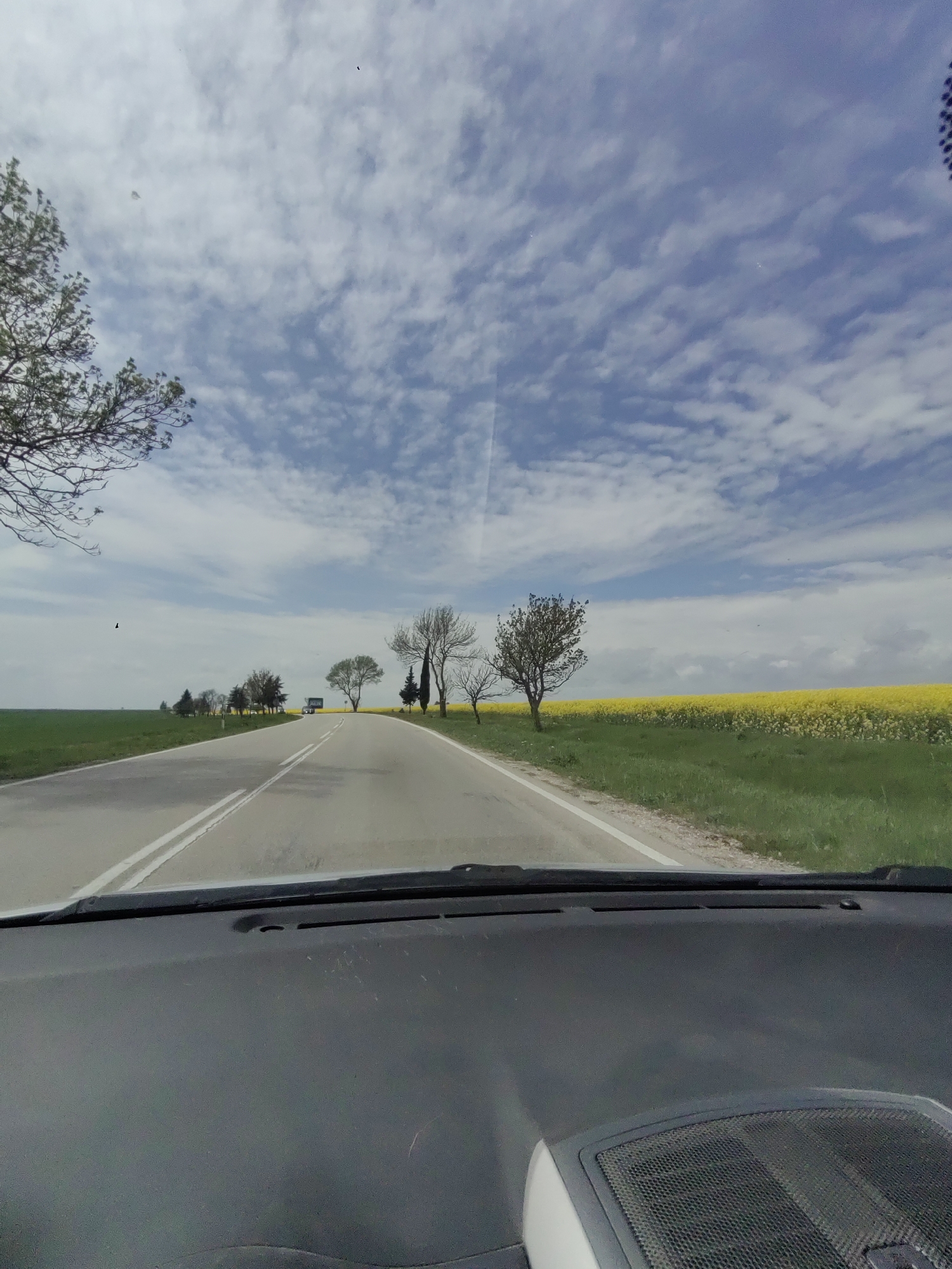 The image shows a view from inside a car, looking out onto a road in a rural area. The car's dashboard is visible in the foreground. The road stretches into the distance, lined with green grass and yellow fields. Trees are sparsely scattered along the roadside. The sky is partly cloudy. It is likely that the location is near Izvorsko, Bulgaria.