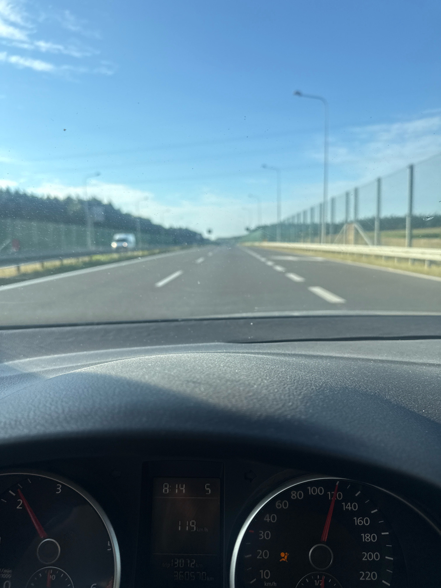 A first-person view from inside a car driving on a highway under a clear blue sky with scattered clouds. The dashboard shows a speedometer with the needle pointing to 120 km/h, and a digital display indicating the time as 8:14 and a speed of 119 km/h, with trip distance at 13072 km and odometer at 360570 km. The road is asphalt with white lane markings, bordered by a green grassy shoulder and a tall fence that appears to be a noise barrier. Trees line the left side of the highway, and a white van is visible in the distance in the oncoming lane. The scene depicts travel on a highway in Popowo, Poland, under daylight conditions.