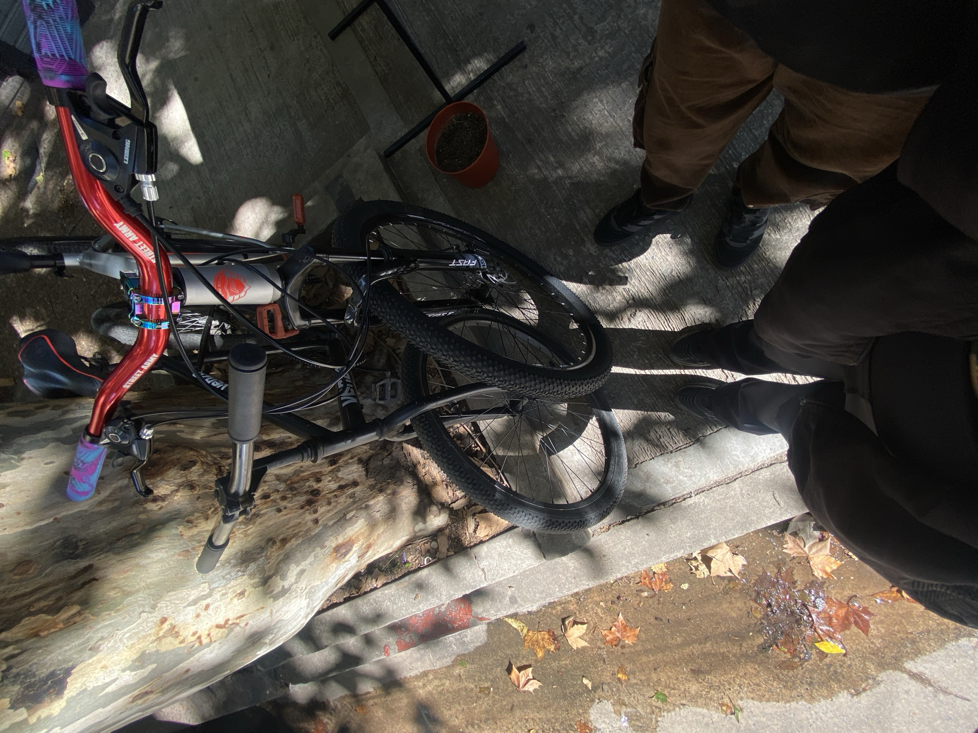 The image presents an outdoor scene in Monte Grande, Argentina, under bright daytime sun, indicated by strong shadows. A bicycle lies on its side, resting partially on a large, light-colored wooden log or tree trunk. The ground is paved and scattered with dry, fallen autumn leaves, suggesting a temperate climate during fall.
The bicycle is a prominent subject, featuring a red handlebar stem clearly labeled "STREET ARMY." Its grips are a vibrant purple and blue pattern, and the black brake lever has visible text that appears to be "L-TWOO." The bicycle's frame is grey or silver with a distinct red graphic, possibly an animal head like a fox or wolf, and the word "VENTURA" is partially visible on the seat or frame. The wheels are black, and "FAST" is printed on one of the tires.
Two individuals are partially visible, seen only from the waist down, standing near the bicycle and the log. One person wears brown pants and black sneakers. The other, mostly obscured by shadow in the foreground, wears darker pants and shoes. Near the person in brown pants, there is a small, empty terracotta plant pot and a black metal frame structure on the pavement. No specific activity is clearly depicted beyond standing or observing the scene.