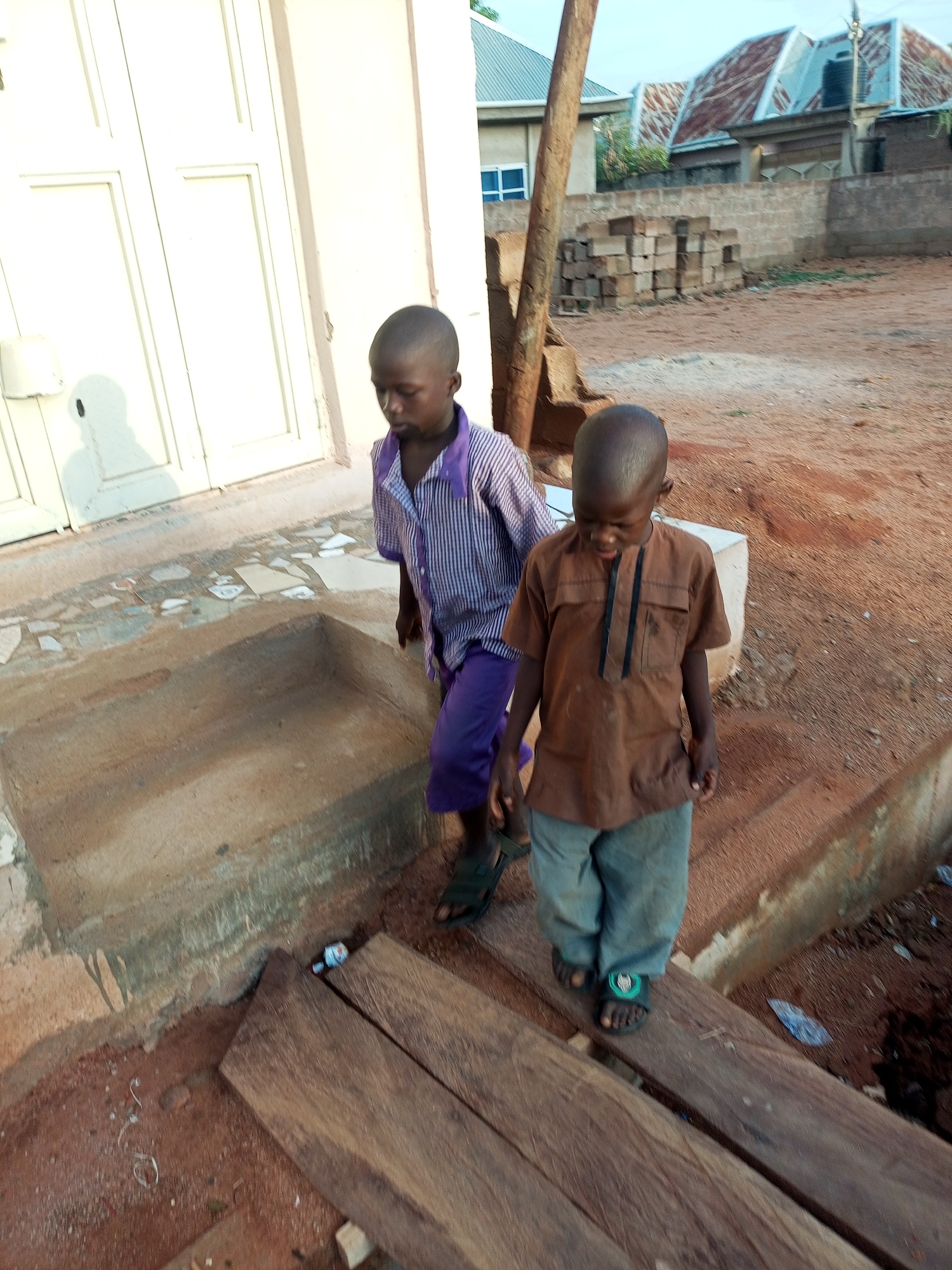 The image features two young boys of African descent walking outdoors in what appears to be a semi-urban or rural setting. The boy on the left wears a purple and white striped short-sleeved shirt with purple shorts or pants and dark sandals. He is positioned slightly behind and to the left of the other boy, looking downward and to his right. The boy on the right wears a short-sleeved brown tunic with black trim down the front, and light blue-grey pants, paired with dark sandals that have a small green emblem on them. He is looking down at his feet or the path ahead.
The boys are traversing an area with reddish-brown earth. In the immediate foreground, two thick wooden planks are laid across a shallow ditch or gap, serving as a makeshift pathway. Scattered small pieces of litter, including white paper or plastic, are visible on the ground and planks. To the left, a light-colored building with large, panelled doors or windows is visible. In the background, there are other structures with corrugated metal roofs, and a stack of concrete building blocks. A bare wooden pole stands vertically on the right side of the image. The lighting suggests either early morning or late afternoon, characterized by soft, warm tones and visible shadows, such as a long shadow cast on the white building to the left. No discernible text is visible in the image.