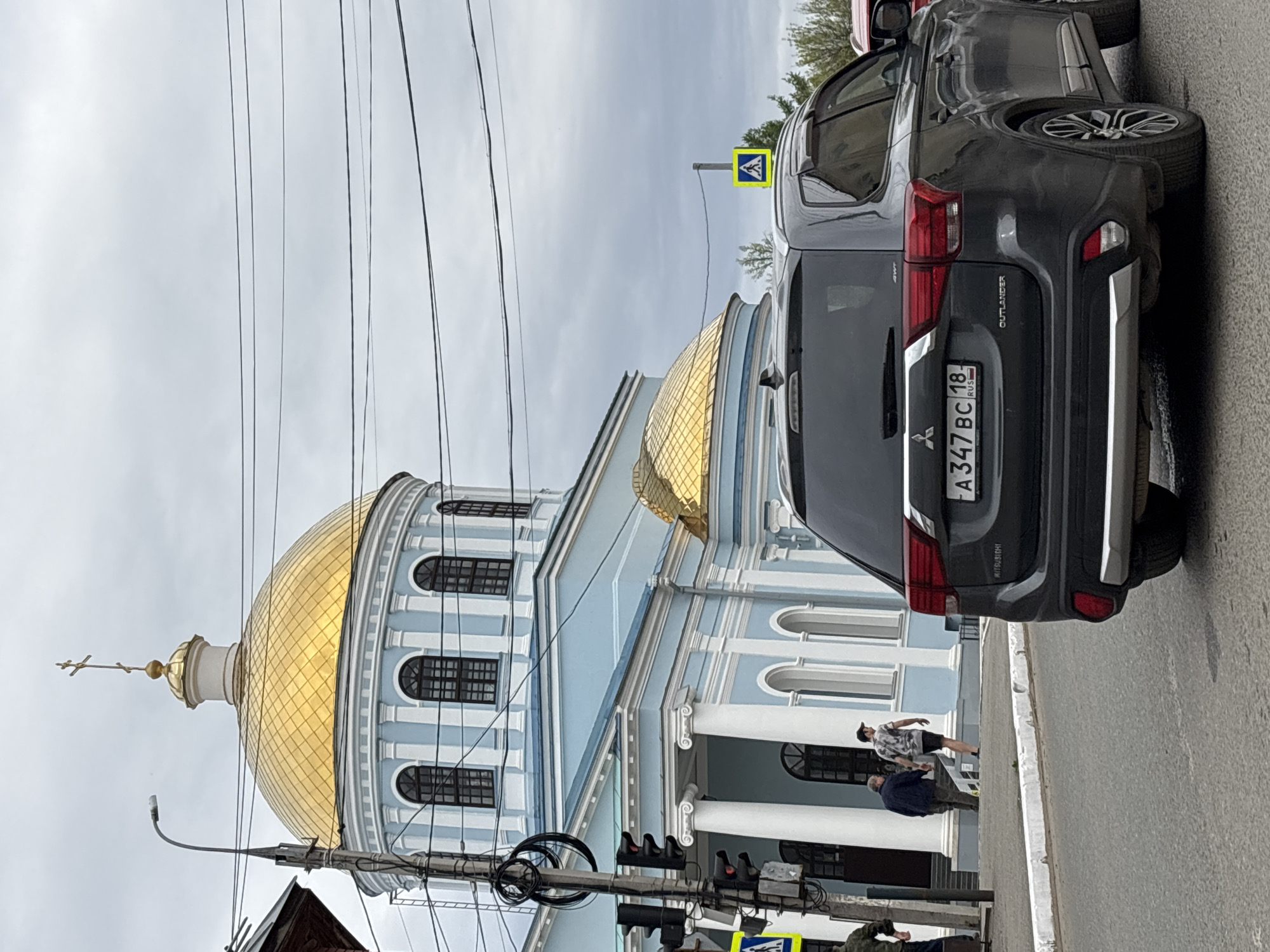 The image depicts a street scene in Votkinsk, Russia, dominated by the Annunciation Cathedral with its large golden dome. In the foreground, a gray Mitsubishi Outlander is parked, its license plate reading "A347 BC 18 RUS." A pedestrian crossing sign is visible in the background, near the vehicle. Several people are walking on the sidewalk near the entrance to the cathedral. Electrical lines crisscross the sky above the building. A traffic light is seen lower in the frame, by the pedestrian crossing, alongside the cathedral.