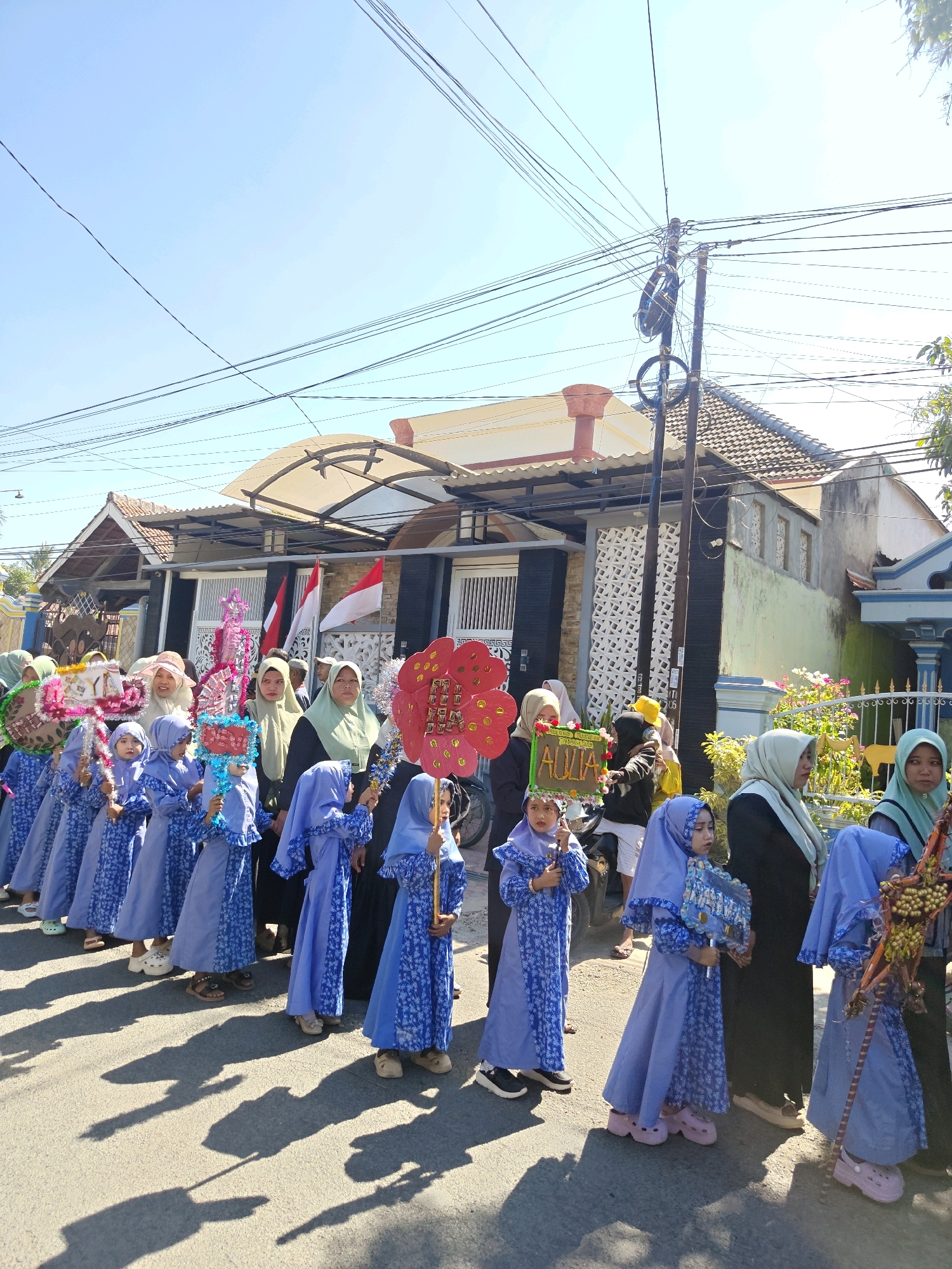 In Klampokan, Indonesia, a procession of young girls dressed in matching blue Islamic attire marches down a paved street under a bright, clear sky. The girls are adorned with colorful headscarves and some carry decorated objects. Several adult women, also wearing headscarves, accompany them, some holding larger decorative items or flags. Behind the procession, residential buildings with tiled roofs and decorative facades line the street. Electrical wires crisscross overhead, strung between poles. The scene captures a community event, likely a celebration or parade.