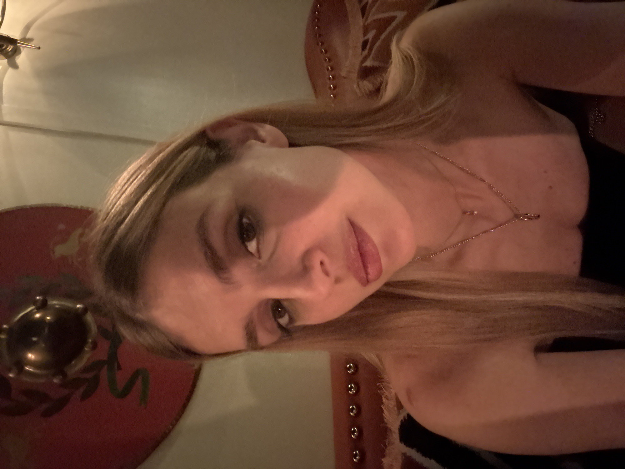 A blonde woman with fair skin is indoors in a building in Tbilisi, Georgia. She has a gold necklace and is wearing a black strapless dress. A circular red decorative piece is visible in the background.