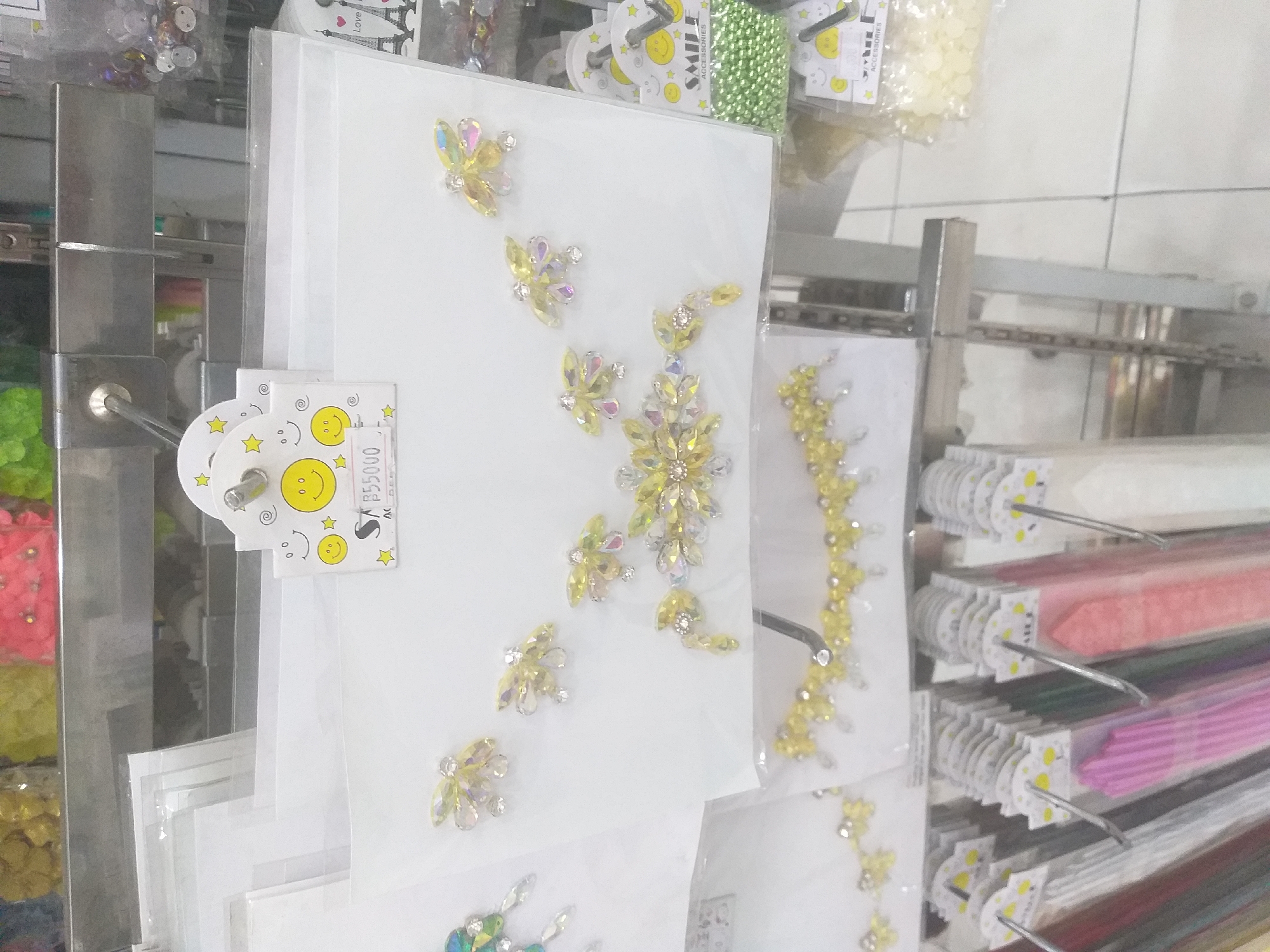 The image depicts a retail display in Batang Serosa, Indonesia, featuring various craft and accessory items. In the foreground, a shelf displays rows of colorful, rolled paper or fabric, likely for crafting. Above this, a white card holds an assortment of sparkly, yellow and clear crystal embellishments arranged in floral and butterfly-like designs. A price tag reading "55000" is visible on the card. To the right, a small bag of green beads hangs, with another package of yellow beads below it, both featuring a "Smile Accessories" label with cartoon smiley faces. The background reveals further shelves with more craft supplies, including what appear to be jars of colorful beads or glitter at the top. The overall scene is consistent with a small shop or market stall specializing in craft materials.