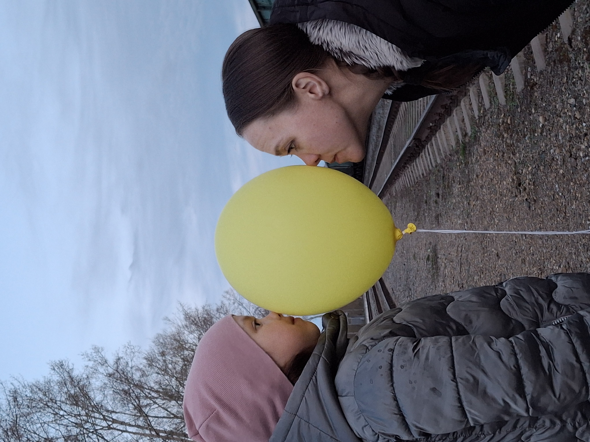 The image, oriented vertically, captures a tender moment between an adult woman and a child, likely a girl, interacting with a bright yellow balloon outdoors. Both individuals are gently pressing their faces against the balloon, creating a sense of closeness and playfulness.
The adult woman, with dark hair pulled back, wears a dark jacket with a white fur-lined hood. Her eyes are subtly closed as she leans towards the balloon. The child, dressed in a pink hooded top and a dark grey puffer jacket, looks intently at the balloon or upwards towards the woman. Their warm clothing suggests cool or cold weather conditions.
The setting is outdoors under a light blue, possibly overcast, sky. Bare tree branches are visible on the left side of the frame, hinting at late autumn, winter, or early spring. On the right, a set of railway tracks with gravel ballast and concrete sleepers stretches into the background, placing the scene near a railway line. The soft, natural light indicates it is daytime. The overall atmosphere is calm and intimate. The location context is Chaikovsky, Russia. There is no visible text in the image.
