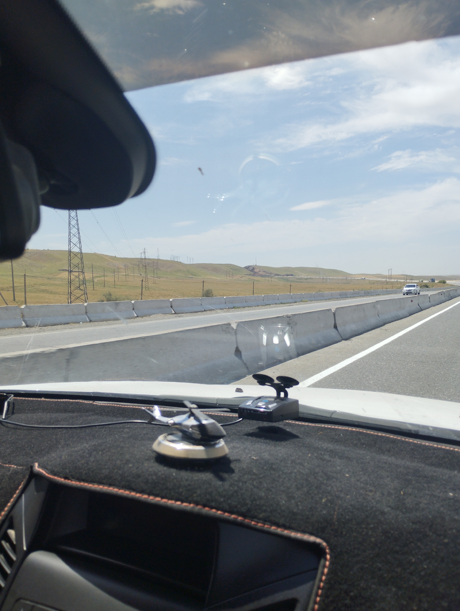 The image, taken from inside a vehicle, depicts a highway scene in Sangzar, Uzbekistan. The foreground shows the dashboard of the car with a black fabric cover and orange stitching. Mounted on the dashboard are two accessories: a black radar detector with suction cups and a small, silver helicopter ornament.
The road ahead is visible, with a concrete barrier separating the lanes. Beyond the barrier, a grassy, hilly landscape stretches to the horizon, dotted with power lines and transmission towers. The sky is partly cloudy, with a bright blue sky and scattered white clouds. In the distance, a white car is seen driving on the highway. The overall impression is one of a clear, sunny day on an open road in a semi-arid rural environment.