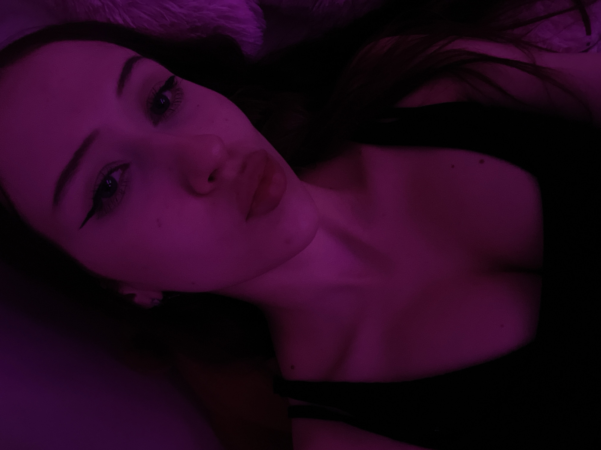 This image is a close-up, overhead shot of a young woman's face and upper body, bathed in purple light. She is looking directly at the camera with her lips slightly parted and her eyes open. Her makeup includes winged eyeliner and mascara. She is wearing a black tank top, and the strap is visible on her left side. The background is dark, with some blurred texture on the right side that appears to be a fluffy material. There is no indication of a specific location within Votkinsk, Russia, or any other environmental details.