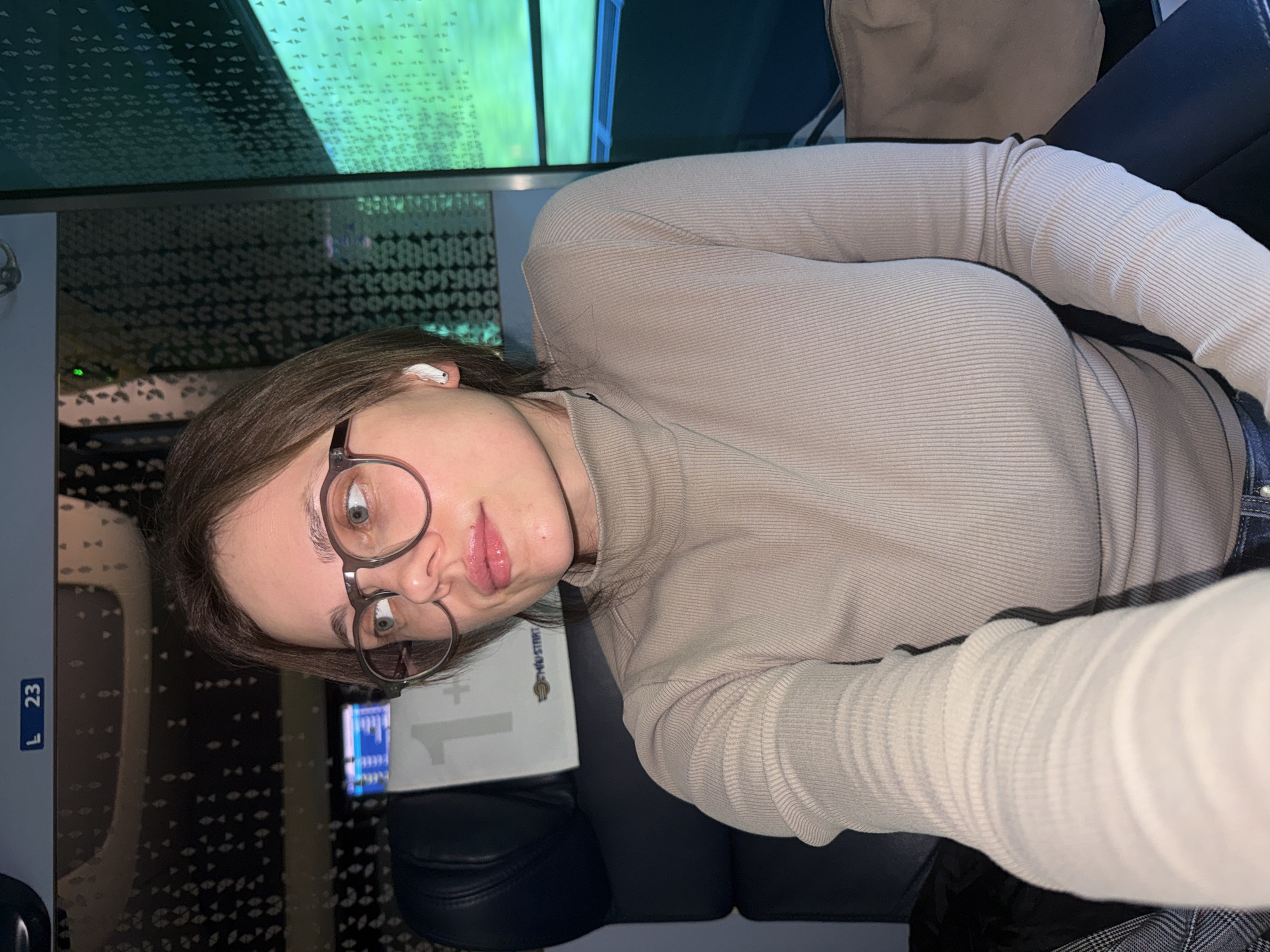 An indoor, portrait-oriented image shows a young woman seated, taking a selfie. She has light brown hair, wears round, dark-rimmed glasses, and a ribbed, light beige long-sleeved top. A white earbud is visible in her right ear. Her gaze is directed towards the camera, with a neutral expression and slightly widened eyes.
The setting appears to be a moving vehicle, possibly a train or bus, indicated by the dark blue seating, a patterned ceiling/wall, and a window displaying a blurred green and blue landscape outside, suggesting motion. The interior lighting is somewhat dim, contrasting with the bright daylight visible through the window.
To her left, a rectangular sign displays "L 23". On a surface next to her, a white card or sign shows a large "1" and a small logo that resembles "YLISTEP" or "TWISTER". The woman seems to be in transit, possibly listening to audio.